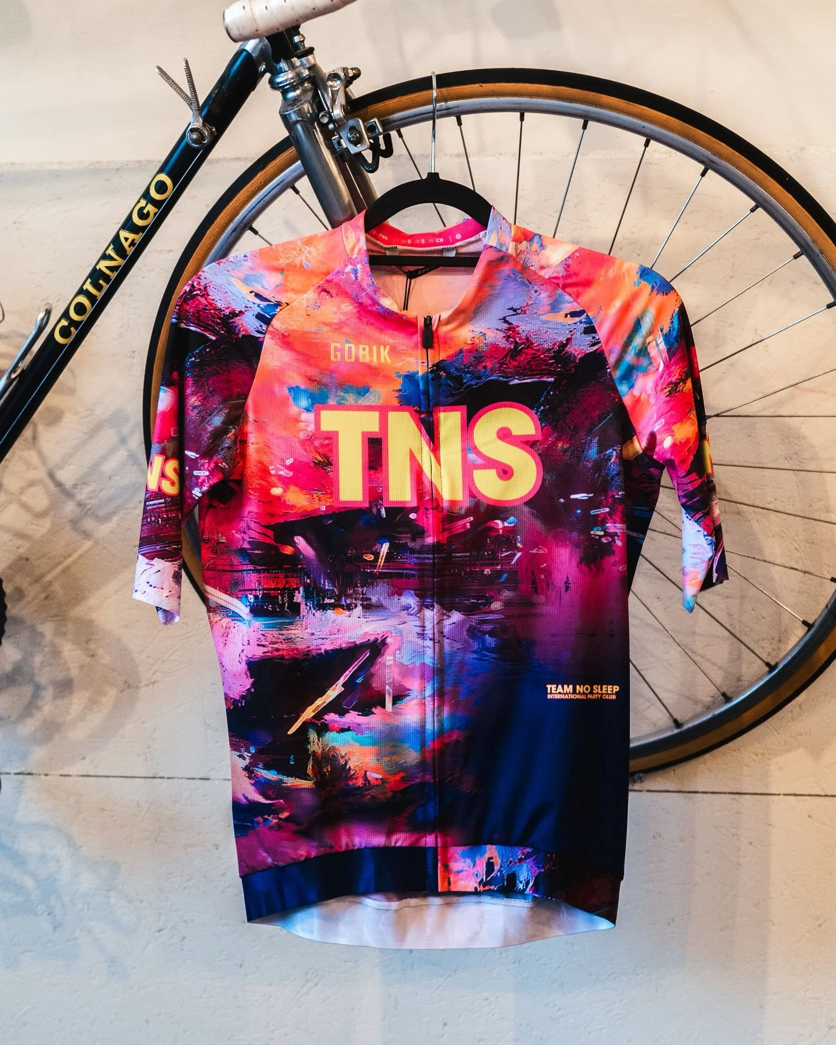 Een kleurrijk wielrentrui met het logo 'TNS' hangt aan een hanger, voor een fiets met racebanden.