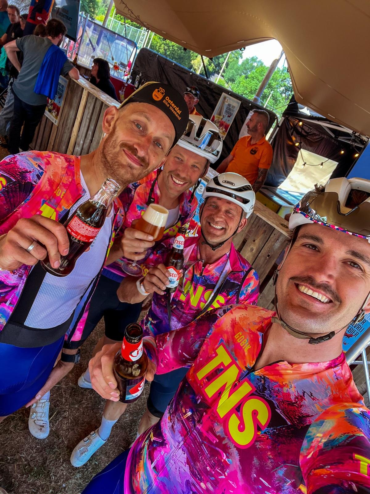 Vier fietsers in kleurrijke shirts en helmen, drinkend en lachend na een fietstocht, onder een tent of schaduwrijke structuur bij een evenement.
