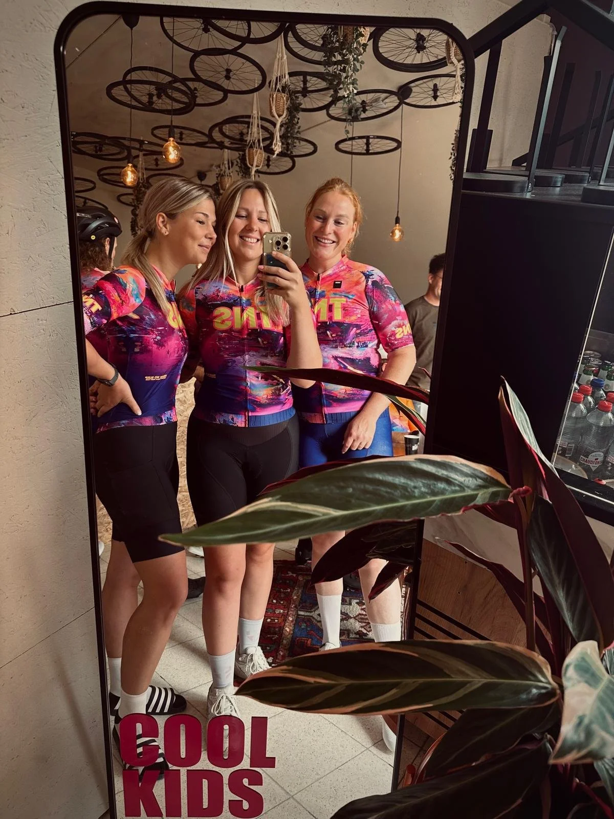 Vijf jonge vrouwen in kleurrijke sportkleding, vier ervan kijken vrolijk naar de spiegel terwijl de vrouw in het midden een foto neemt. Ze staan binnen, onder decoratieve verlichting en planten, met een tekst op de spiegel die zegt 'COOL KIDS'.