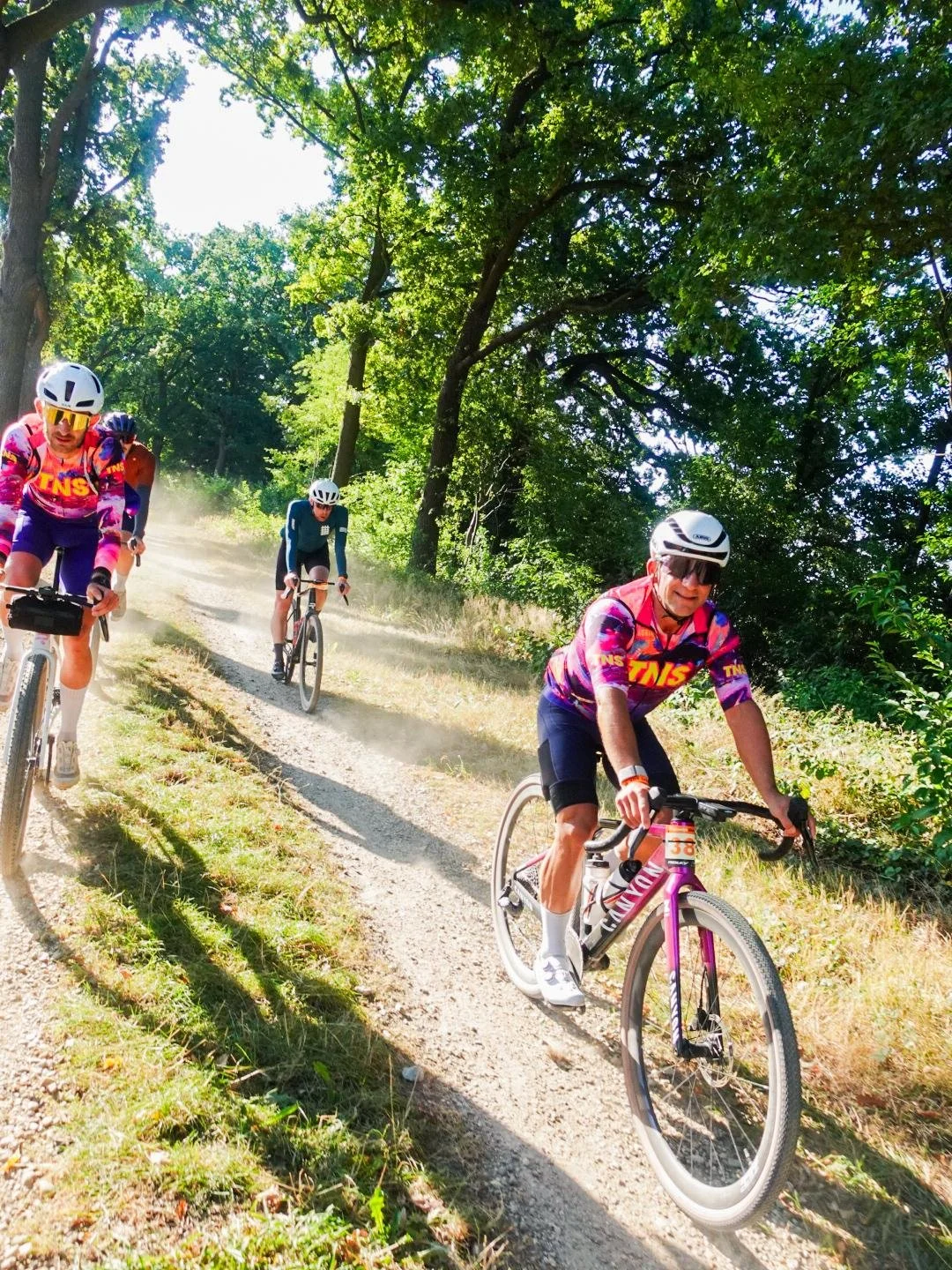 Vijf mountainbikers berijden een stoffig pad door een bos met groene bladeren onder een heldere blauwe hemel.