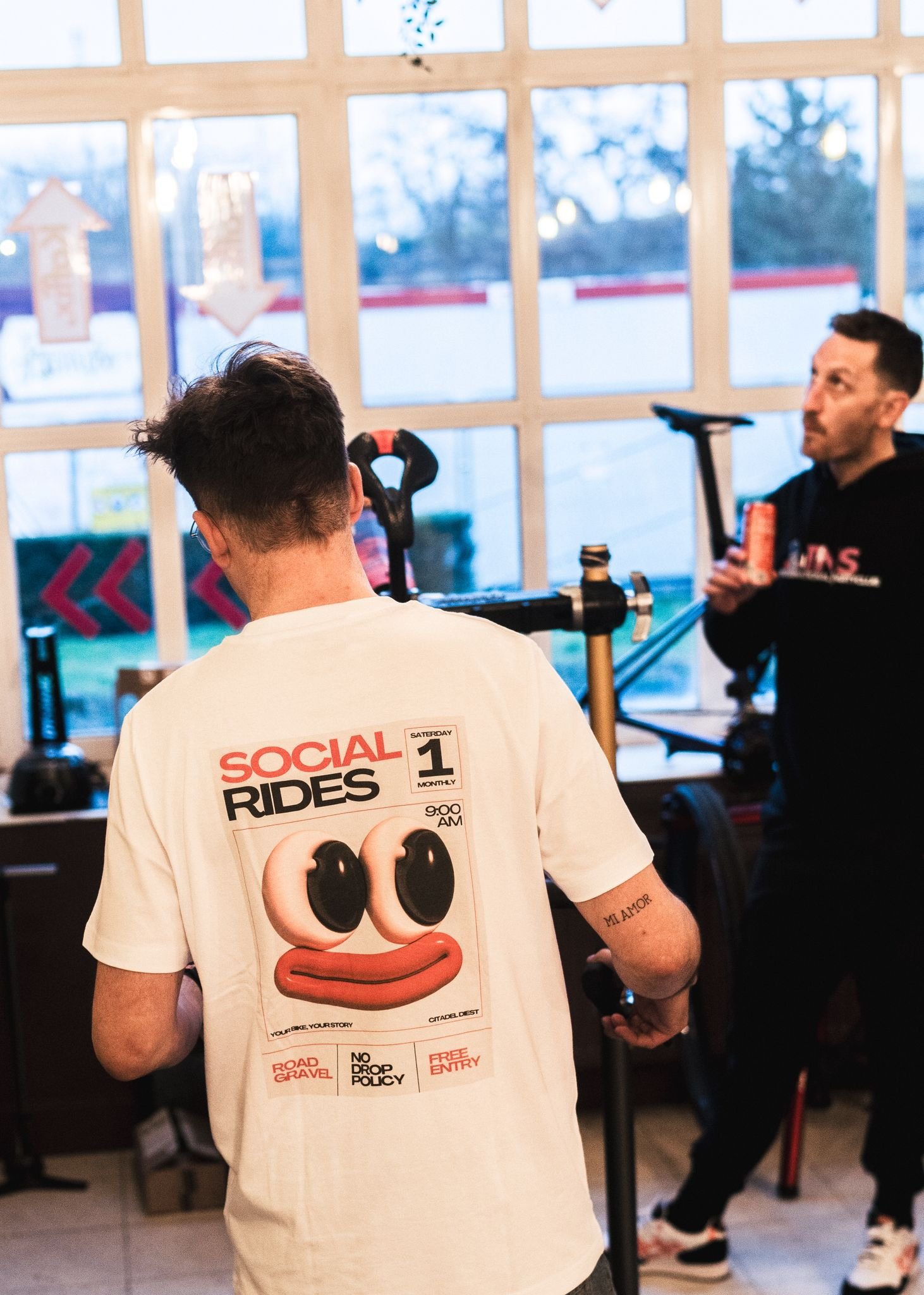 Twee mensen in een fitnessruimte, een van hen met een witte T-shirt met een cartoonachtig gezicht en tekst over sociale ritten, en de ander met een zwart hoodie met rode tekst, in de achtergrond fitnessapparatuur en grote ramen met uitzicht op een wi