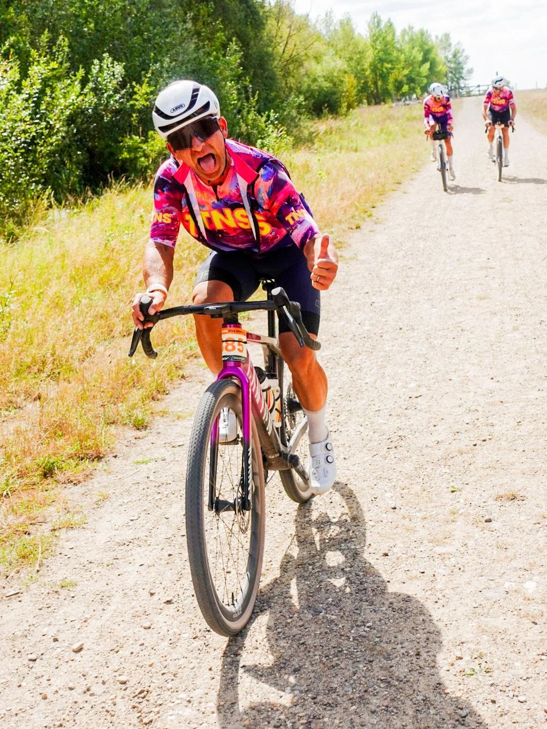Een man fietst op een onverharde weg in de natuur, hij draagt een helm en een kleurrijke wielershirt. Hij geeft een duim omhoog en lijkt te genieten. Twee andere fietsers volgen op de achtergrond.