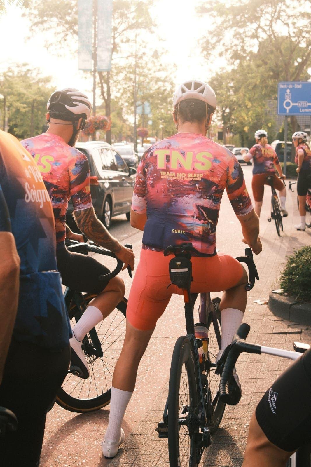 Mannen in kleurrijke fietskleding en helmen wachten op de fiets op een stadsplein, met auto’s en wandelende mensen op de achtergrond.