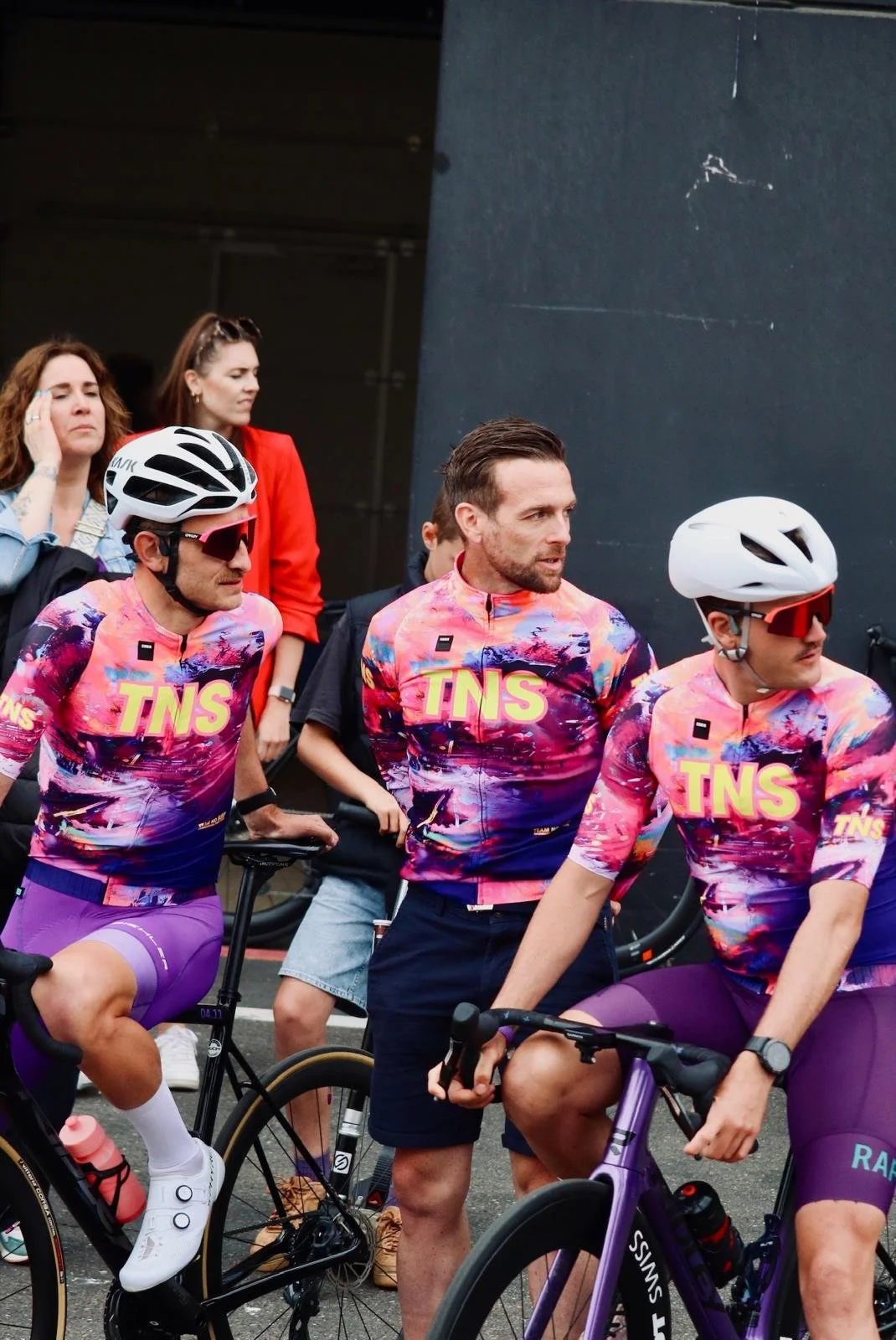 Tre mannen in kleurrijke wieleroutfits met 'TNS' logo, klaar voor een fietswedstrijd.