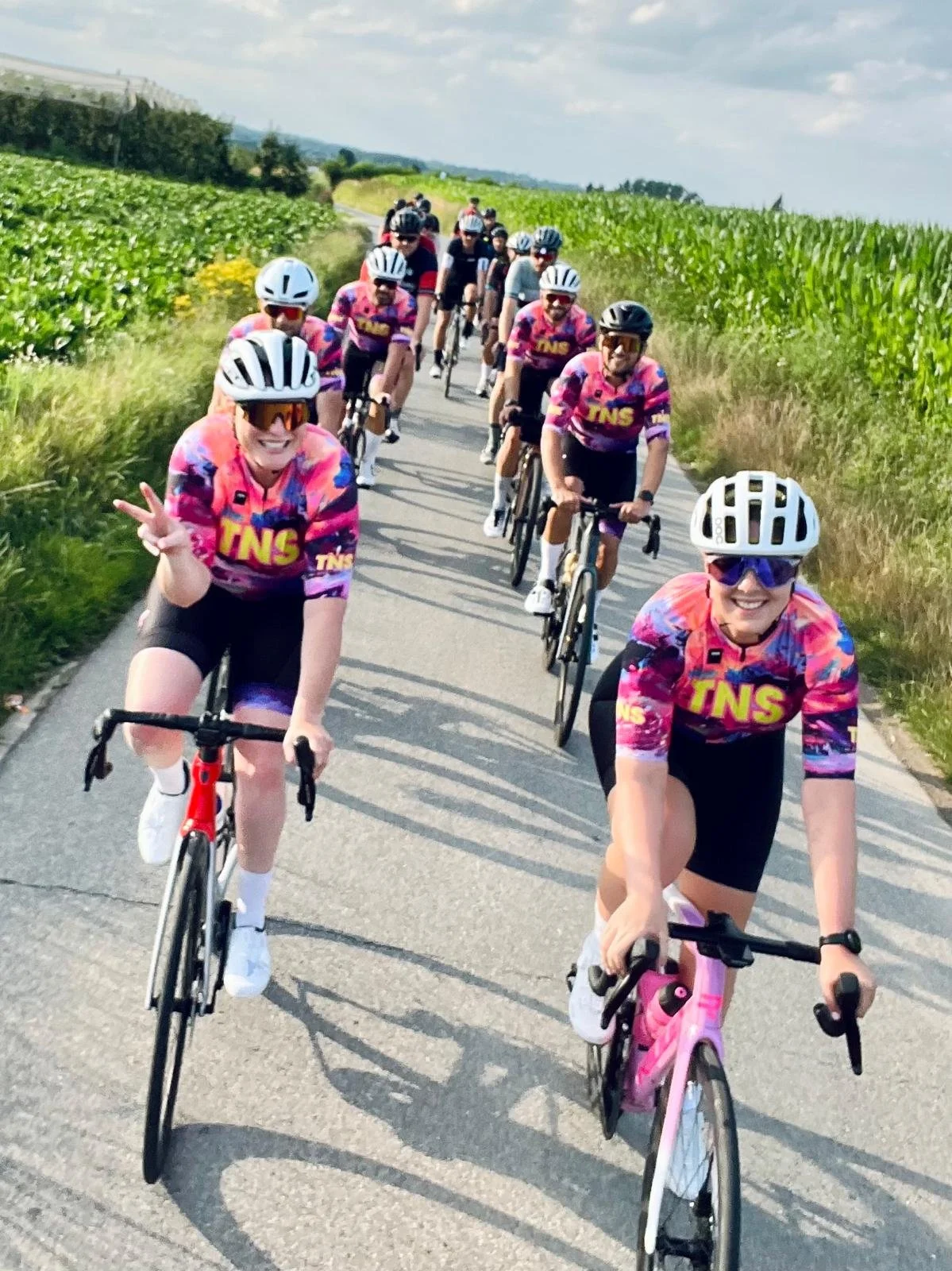 Groep fietsers in kleurrijke shirts rijden op een landweggetje, omgeven door groene velden onder een bewolkte hemel.