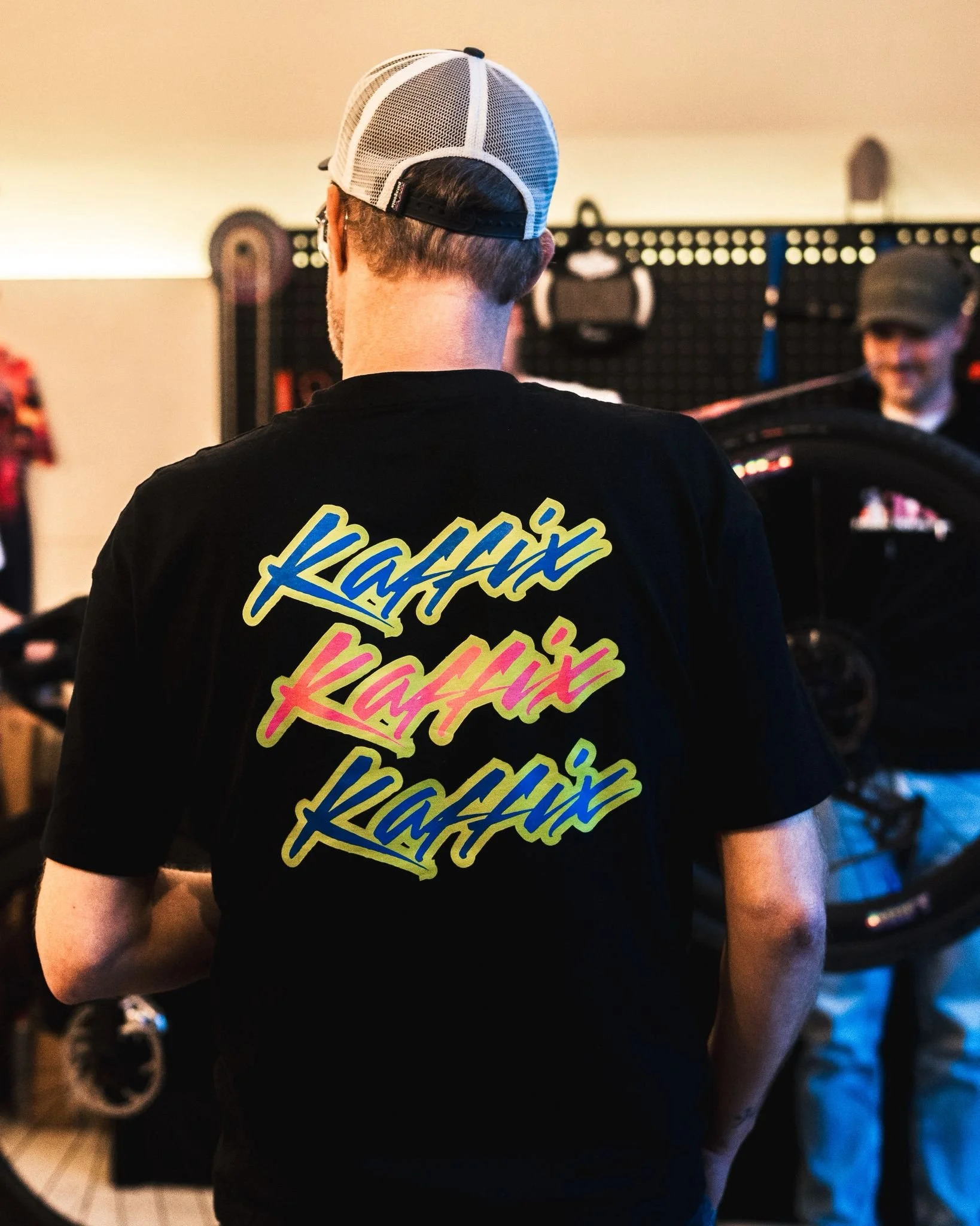 Man met zwart T-shirt met kleurrijke 'RAFFIX' tekst, achterzicht, in een winkel of werkplaats met fiets en gereedschap.