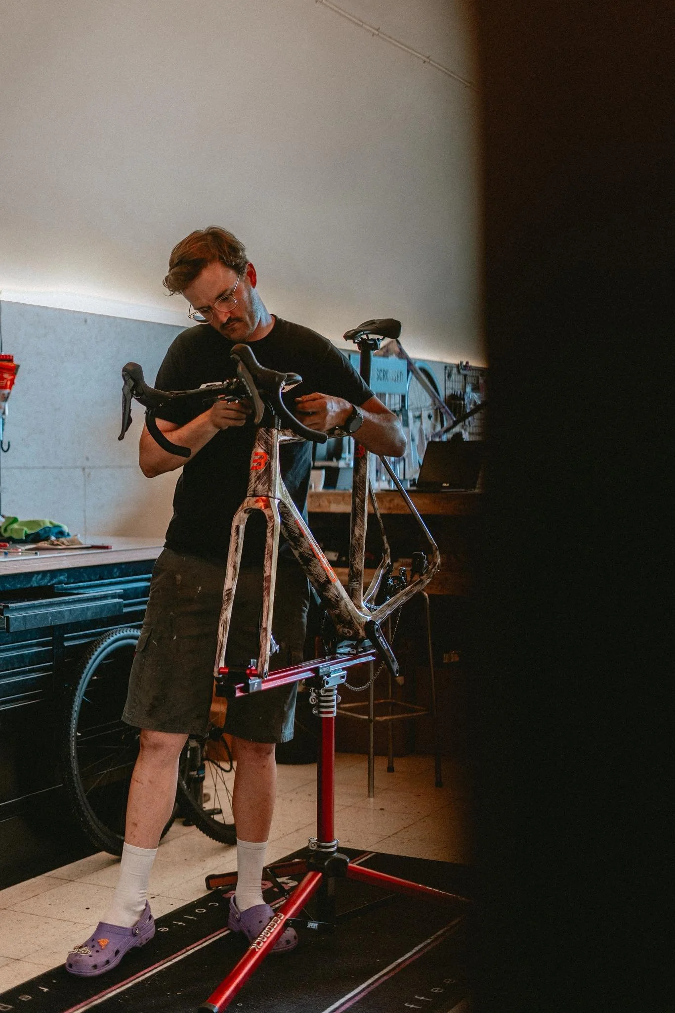 A man working on a bicycle frame in a workshop, standing on a red repair stand, with tools and equipment in the background.