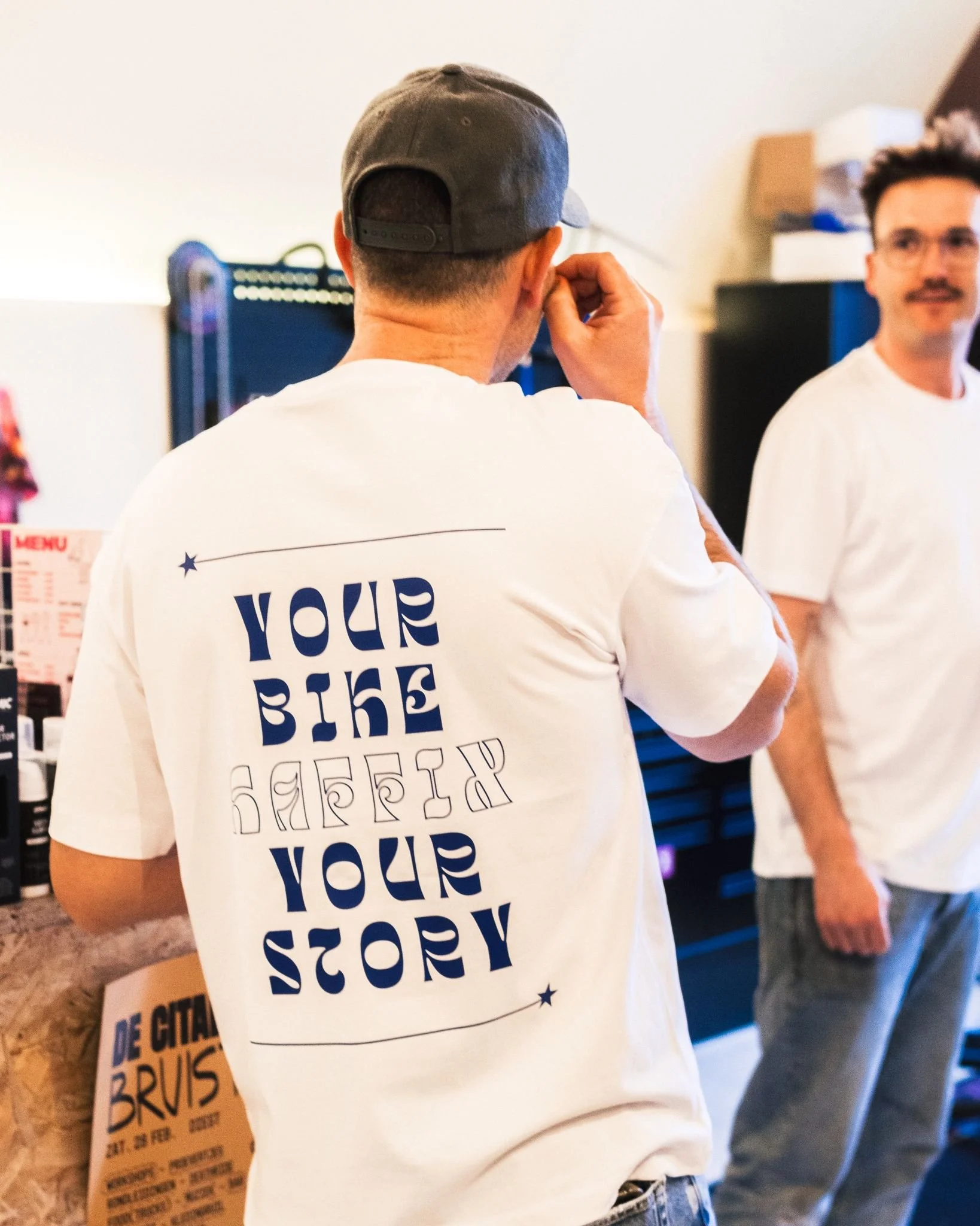 Twee mannen in witte t-shirts, eentje met de achterkant zichtbaar en de ander met een gezicht, in een winkel of café. De achterzijde van de t-shirt heeft de tekst 'YOUR STORY' in grote letters.
