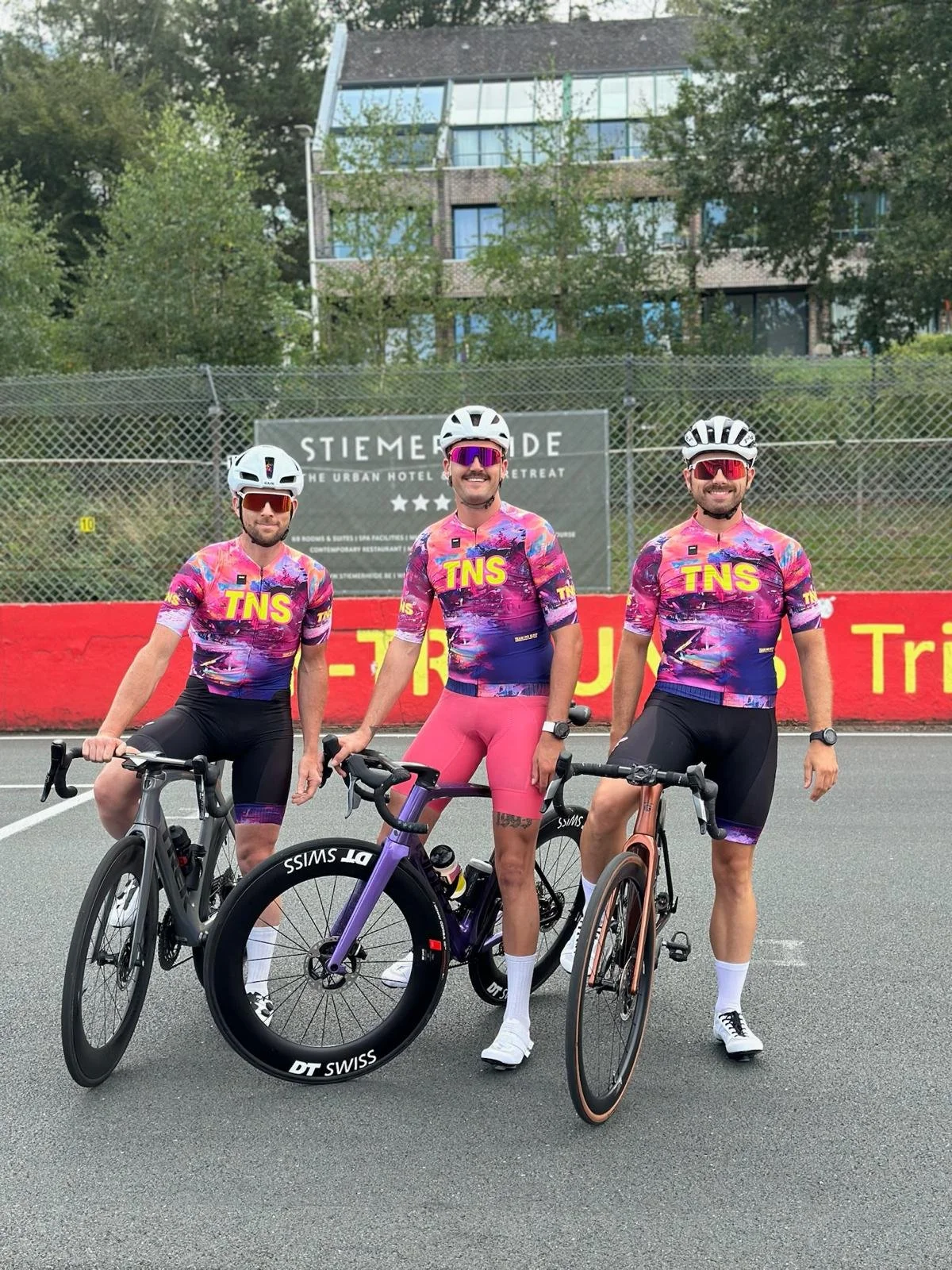 Drie wielrenners in kleurrijke fietskleding staan naast hun fietsen op een weg, met een hek en een gebouw op de achtergrond.