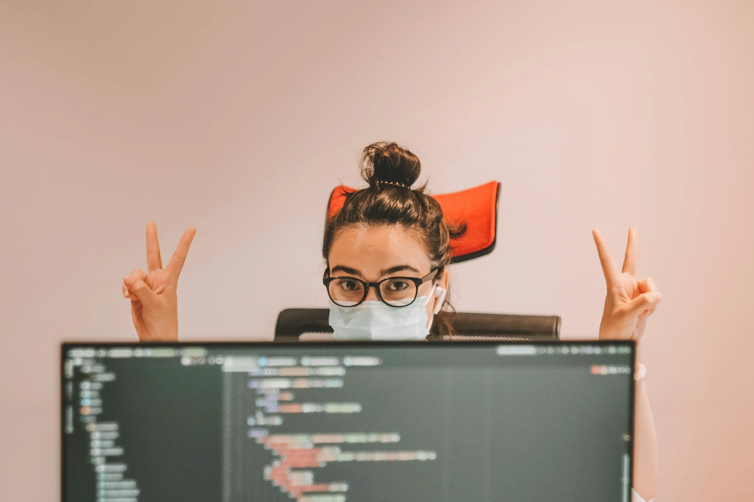 Young woman with glasses and face mask sitting at a desk with a computer screen in front, raising both hands making peace signs, in an office environment.
