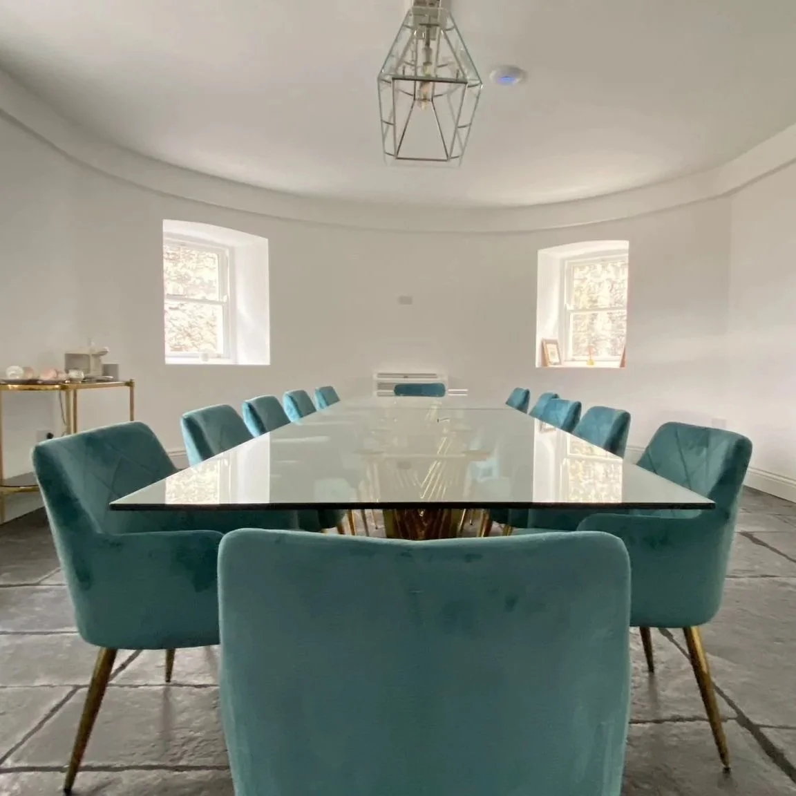 A modern conference room with a large oval glass table surrounded by teal velvet chairs, two windows, and a minimalist geometric pendant light fixture.