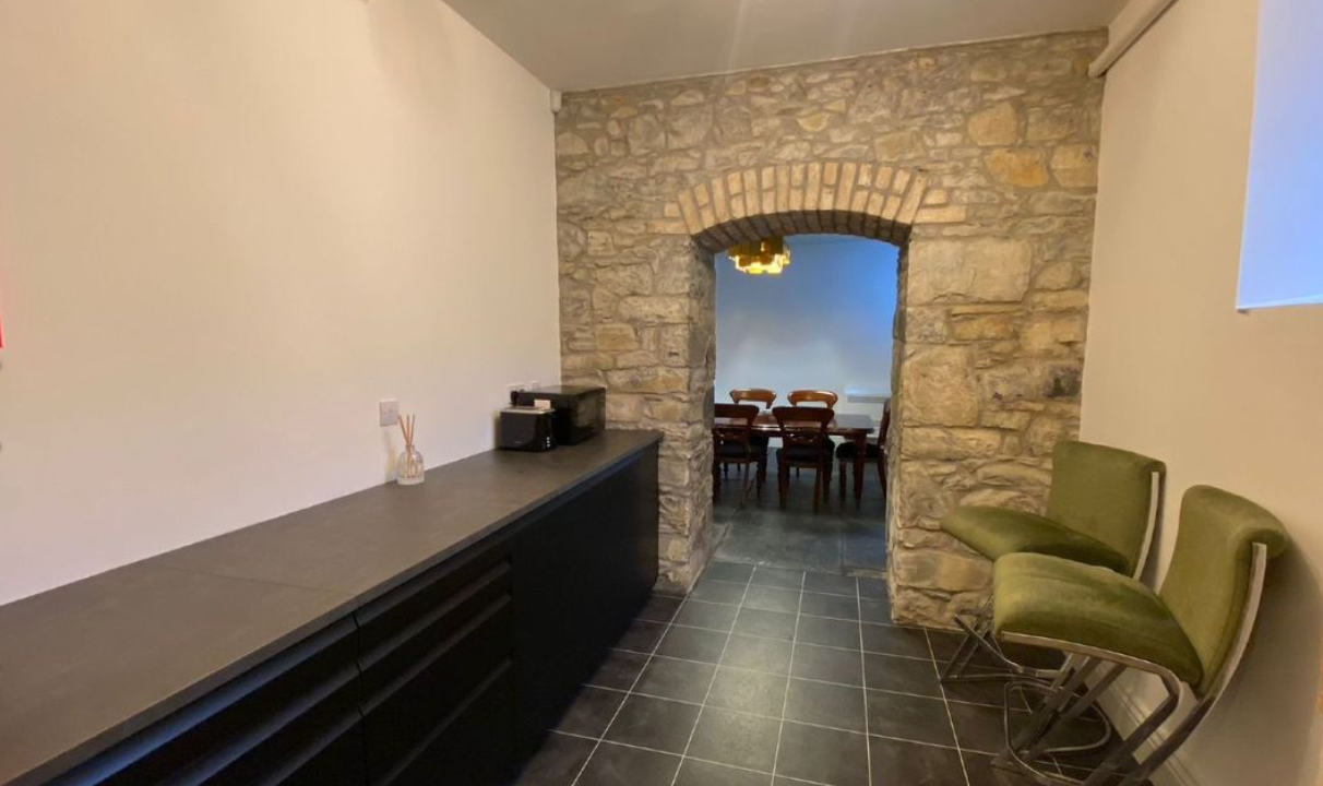 Interior of a room with a stone archway leading to a dining area with a wooden table and chairs. The room has black tiled flooring, a black countertop with some items on it, and two green chairs along the wall.