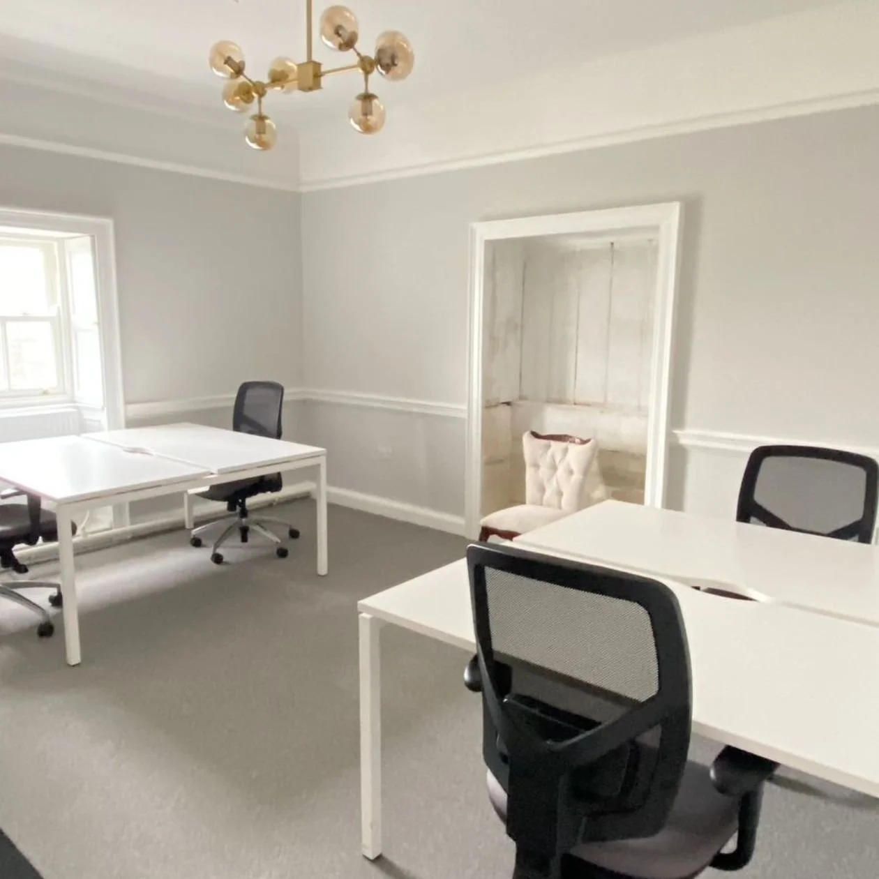 Empty office with white desks, black office chairs, a beige upholstered chair, and a window letting in natural light, with a modern chandelier hanging from the ceiling.