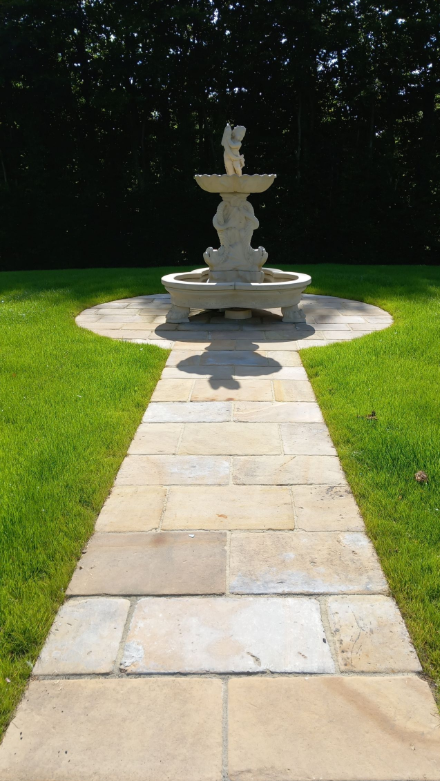 Stone pathway leading to a decorative white fountain with a cherub figure in a grassy yard with trees in the background.