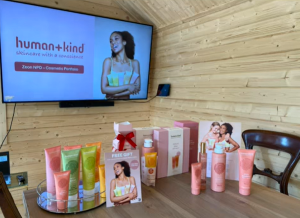 Display of skincare products on a wooden table with promotional material, in a room with wooden walls, including a large screen showcasing a woman and the words 'human+kind skincare with a conscience'.