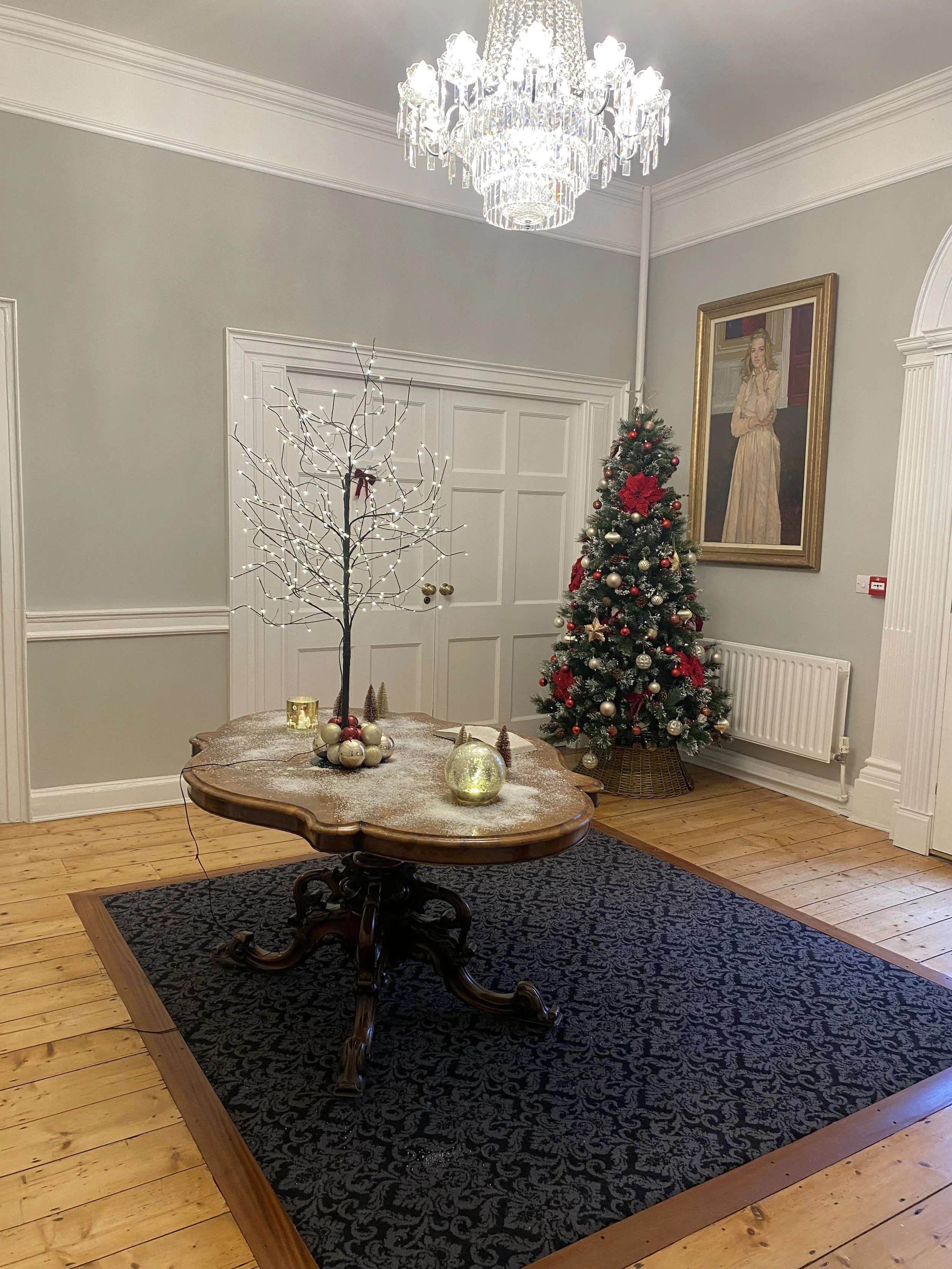 A decorated Christmas tree with red and gold ornaments and poinsettia flowers in a living room. A white illuminated branch centerpiece sits on a wooden table, which is on a black patterned rug. A chandelier hangs from the ceiling, and a framed painti