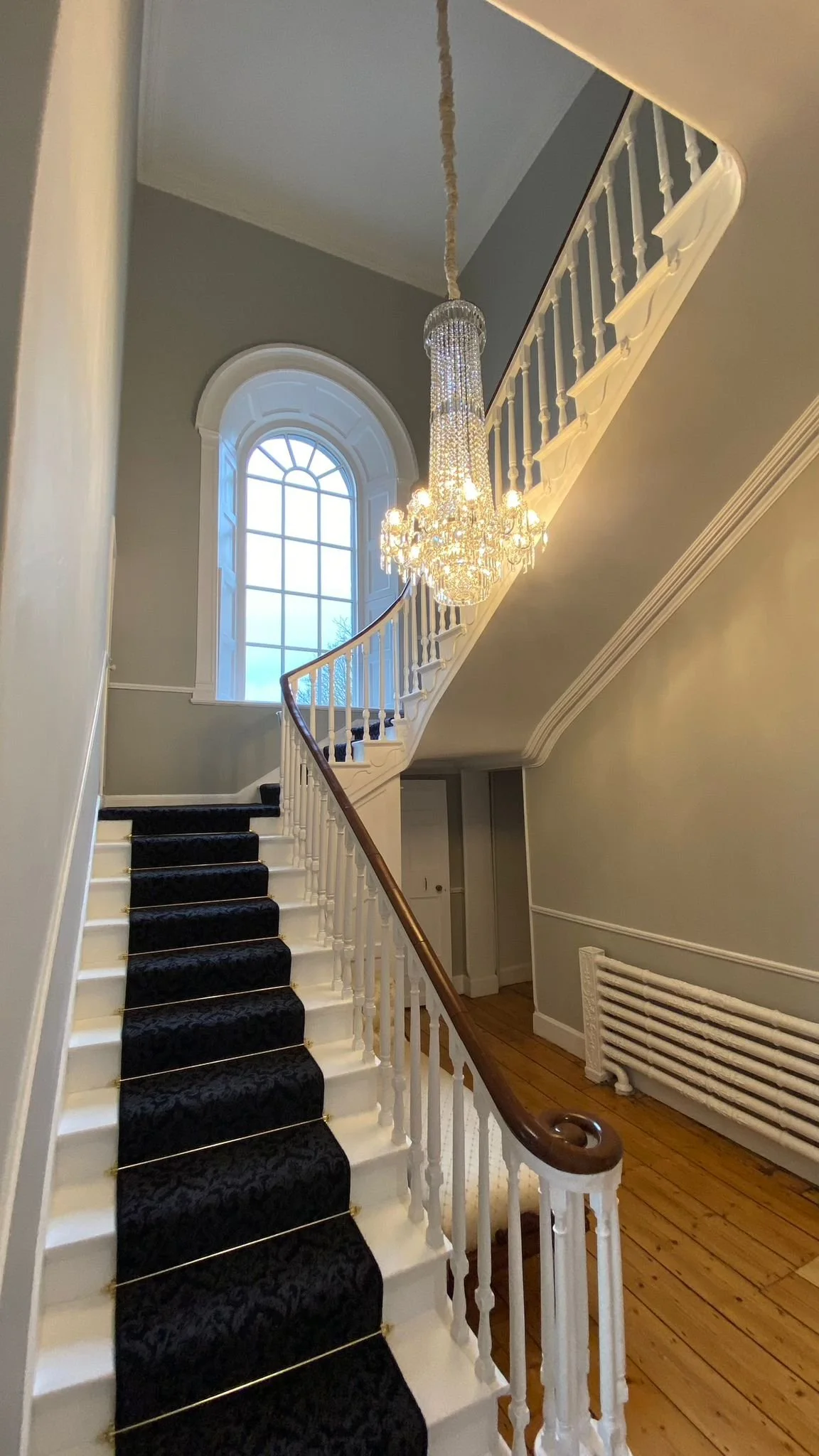 Interior staircase with white balustrade, black carpet runner, wooden handrail, large arched window, and chandelier.