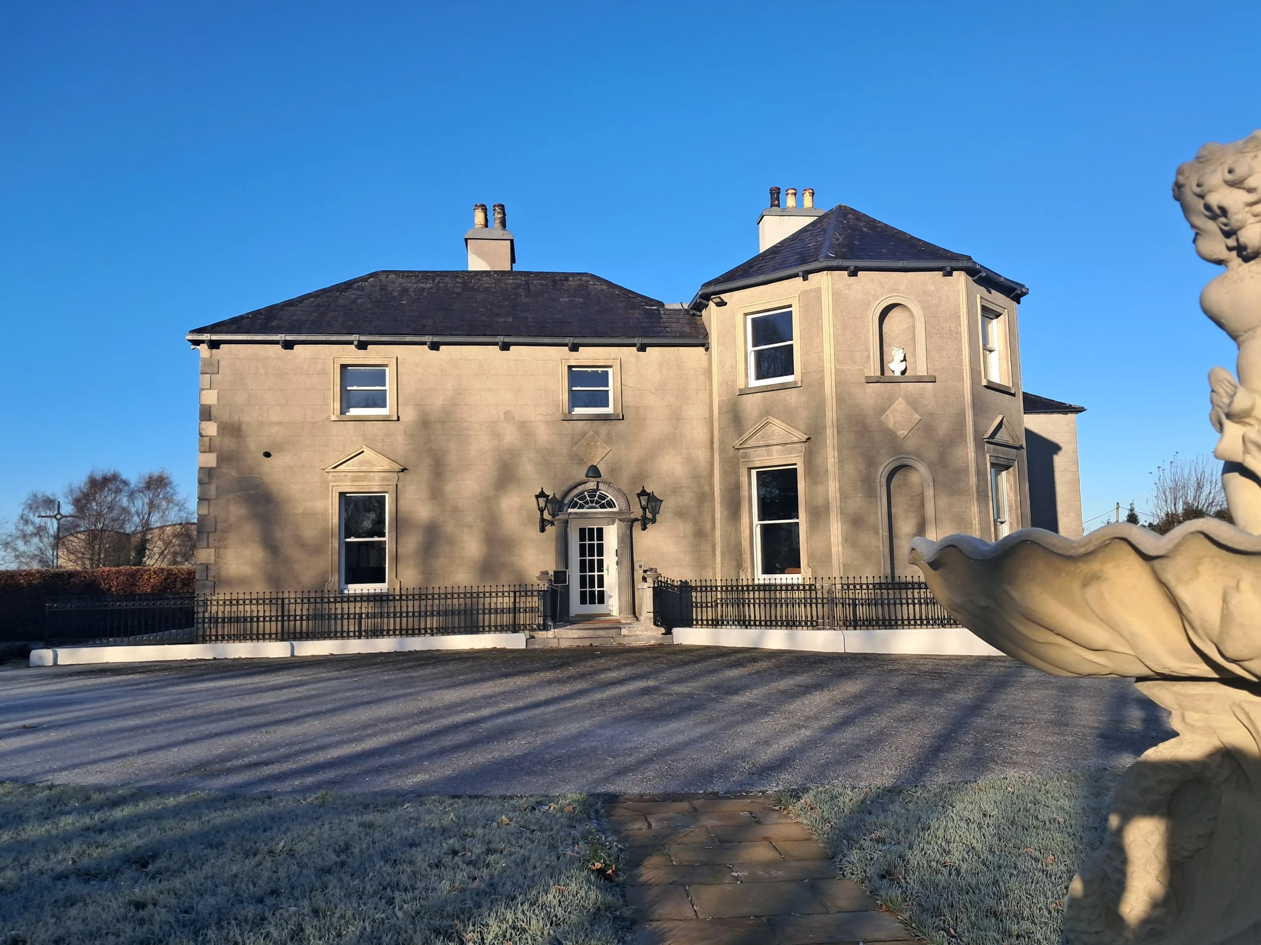 A large, Victorian-style house with beige walls, black roofing, multiple chimneys, and several windows, situated behind a black iron fence. There is a stone fountain on the right side of the image and a gravel driveway in front of the house. The scene is illuminated by sunlight with a clear blue sky overhead.