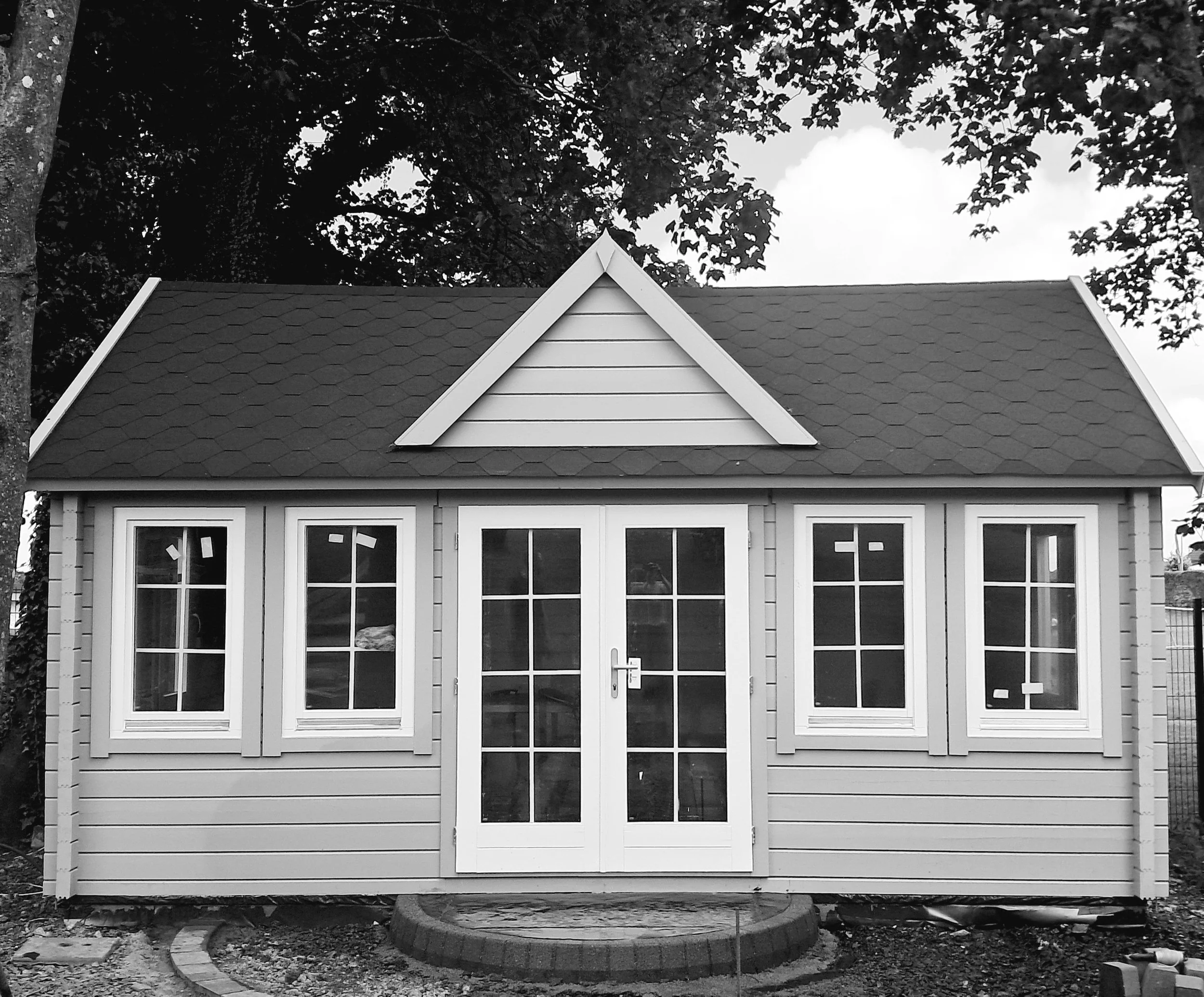 A small house with a triangular gable roof, four windows, and double glass doors, surrounded by trees in the background.