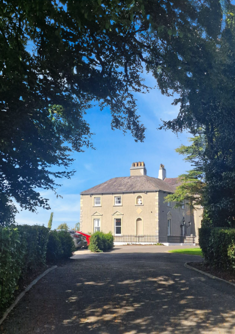 A large, beige, historic house with multiple windows and chimneys, surrounded by trees and greenery, with a curved driveway leading up to it.