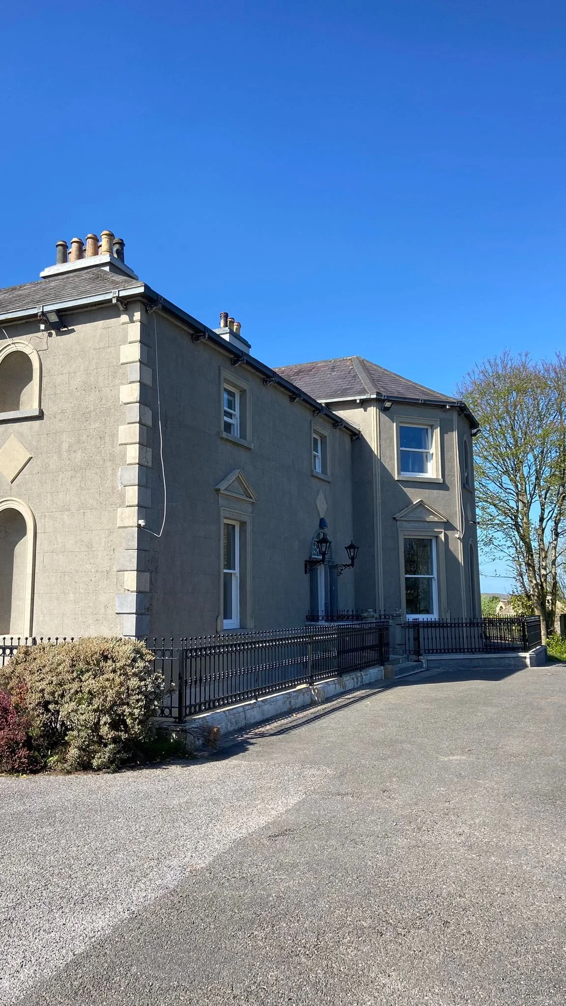 A large gray stone house with multiple windows, black metal fence, and a driveway, under a clear blue sky.
