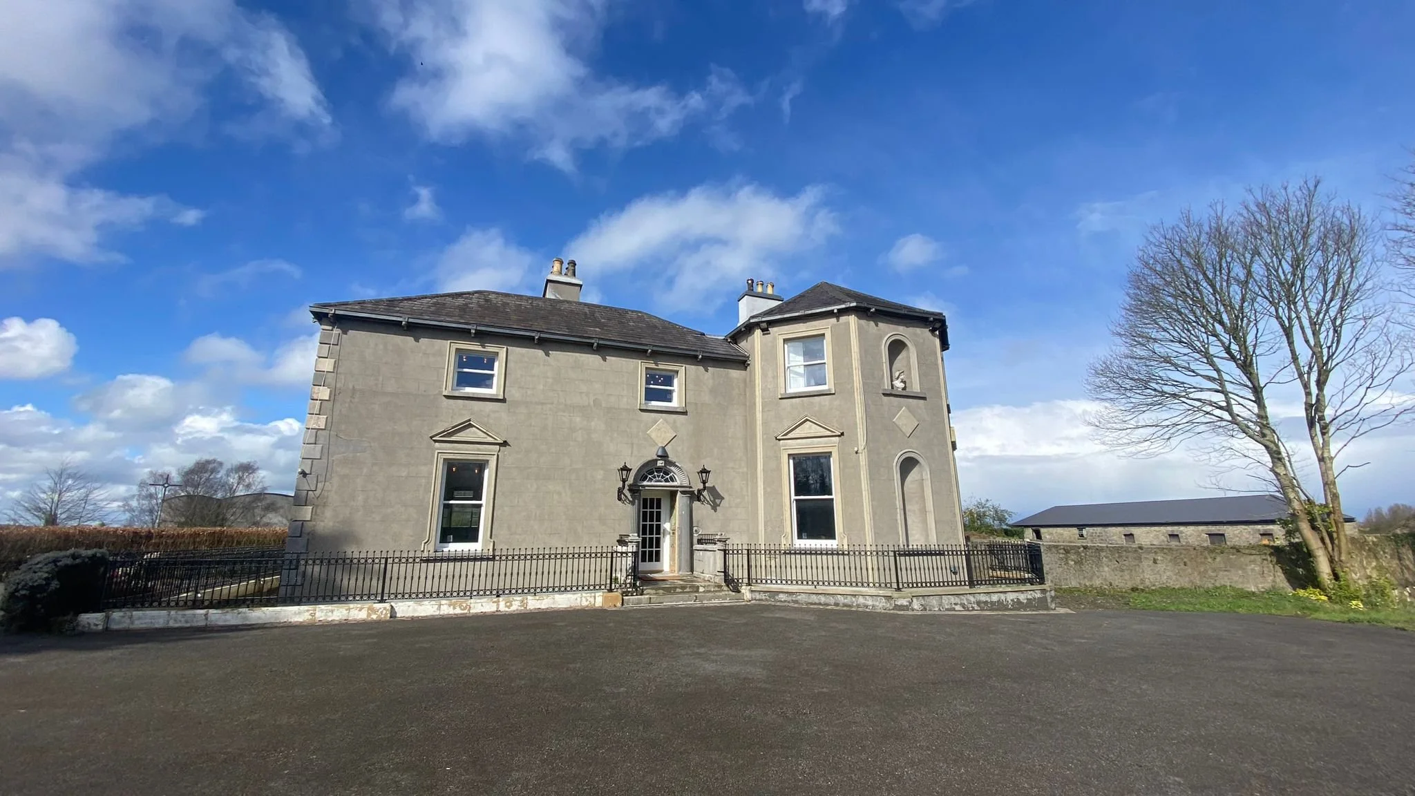 A large, old, gray mansion with decorative architectural features, black iron fence, and a clear blue sky with some clouds.