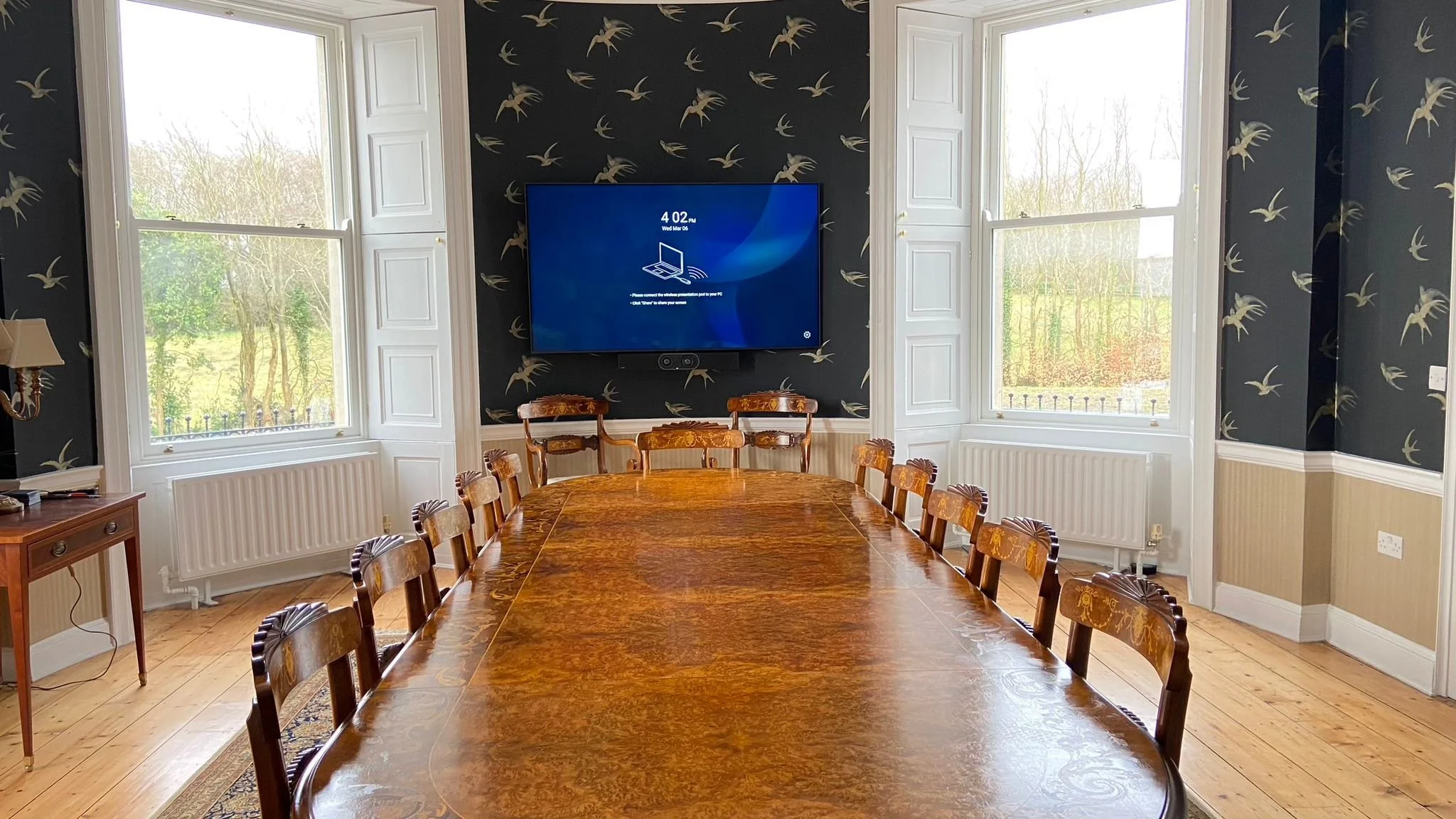 A conference room with a long wooden table and ten chairs, surrounded by three large windows with white frames and plantation shutters. The room has dark wallpaper with a bird pattern, and a wall-mounted TV displaying a screen with the time 4:02 pm a