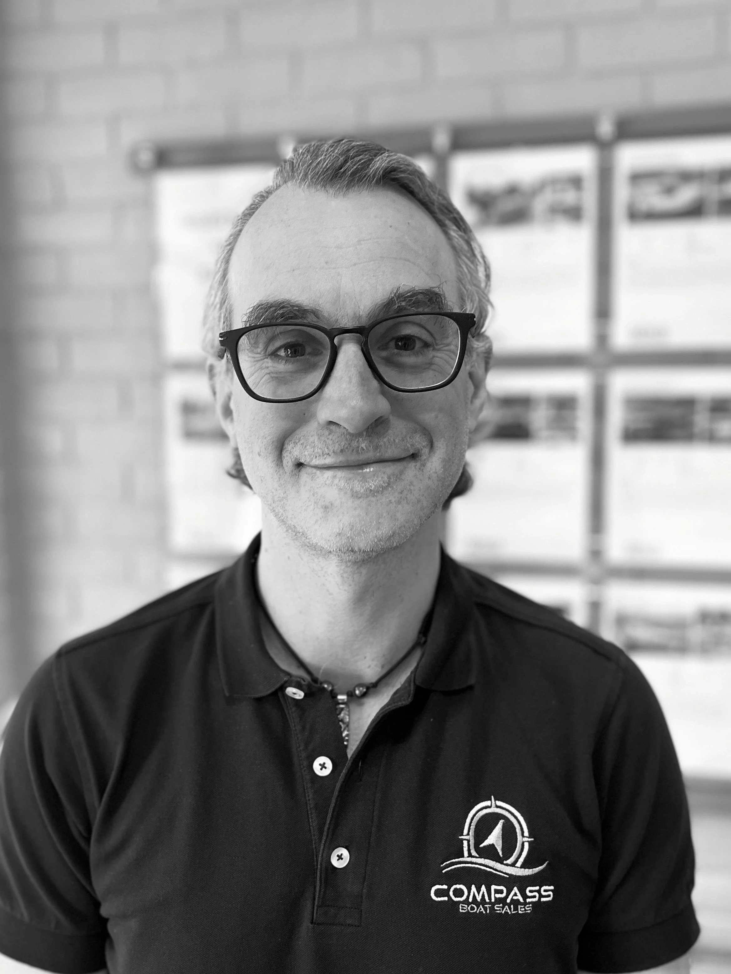 A smiling man wearing glasses and a black polo shirt with a 'Compass Boat Sales' logo, standing in front of a brick wall with pictures.