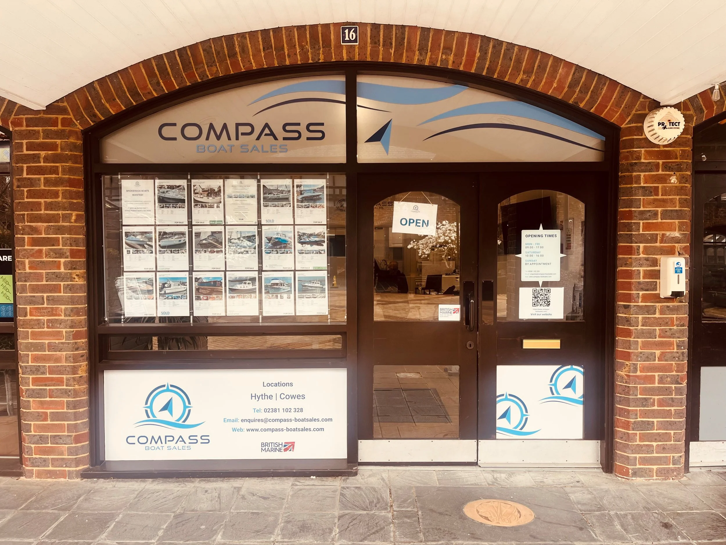 The storefront of Compass Boat Sales, with a large window display of boat listings and a glass door marked "Open," showing the store's logo, location details, contact information, and signage about opening times.