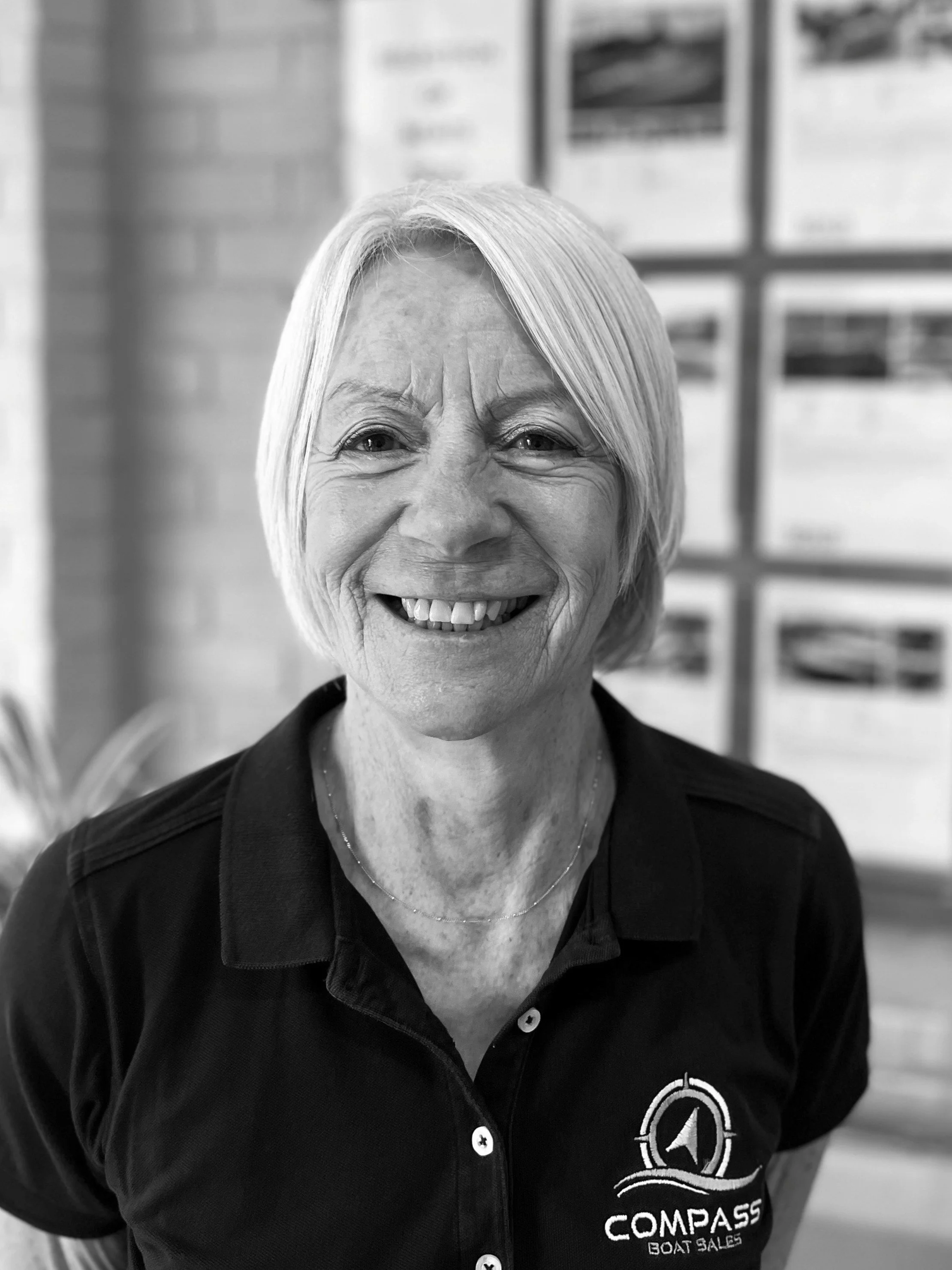 A smiling elderly woman with short, light-colored hair, wearing a black polo shirt with 'COMPASS BOAT SALES' logo, standing indoors.
