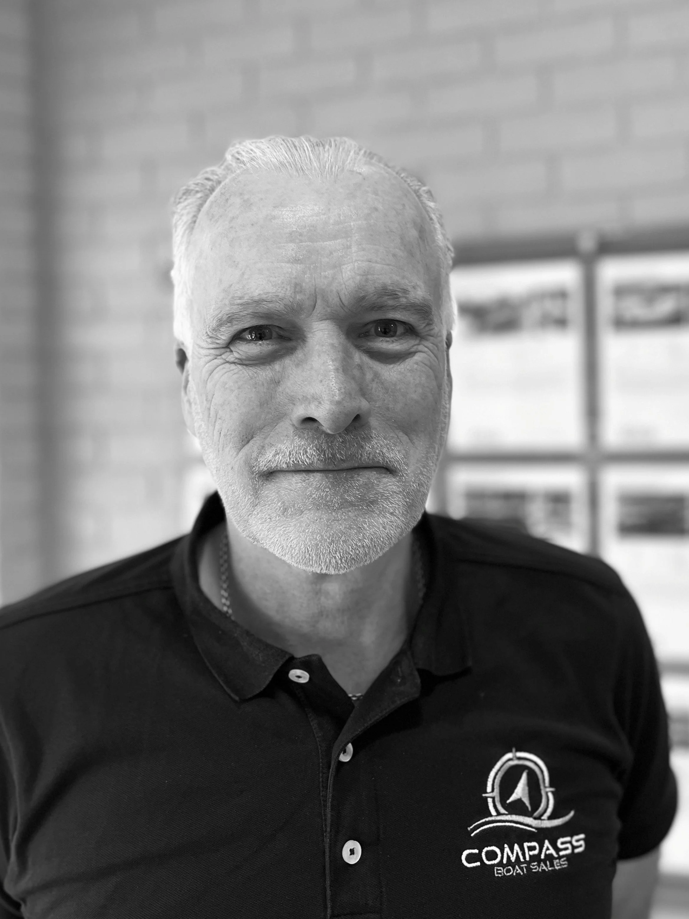 A middle-aged man with light hair and a beard, wearing a black polo shirt with a 'COMPASS BOAT SALES' logo, standing indoors in front of a brick wall and framed pictures.