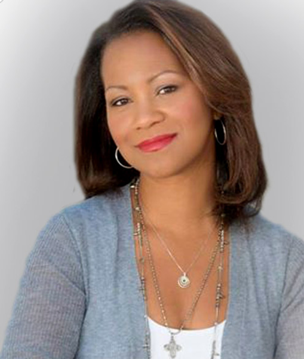 Woman with shoulder-length brown hair, wearing a light gray cardigan over a white top, accessorized with layered necklaces and hoop earrings, smiling softly.