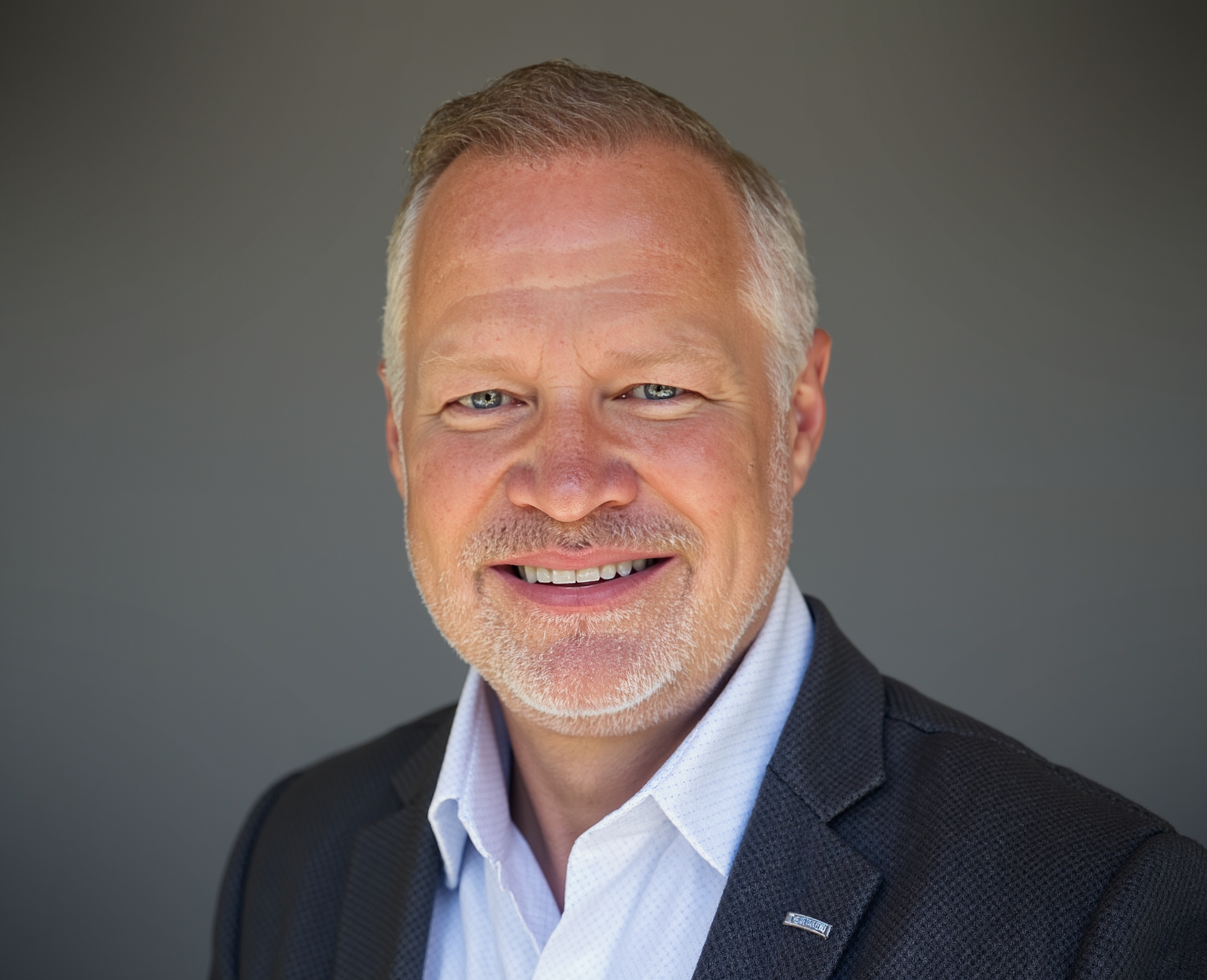 Close-up of a middle-aged man with short gray hair, blue eyes, and a smile, wearing a dark suit and a white shirt, against a plain gray background.