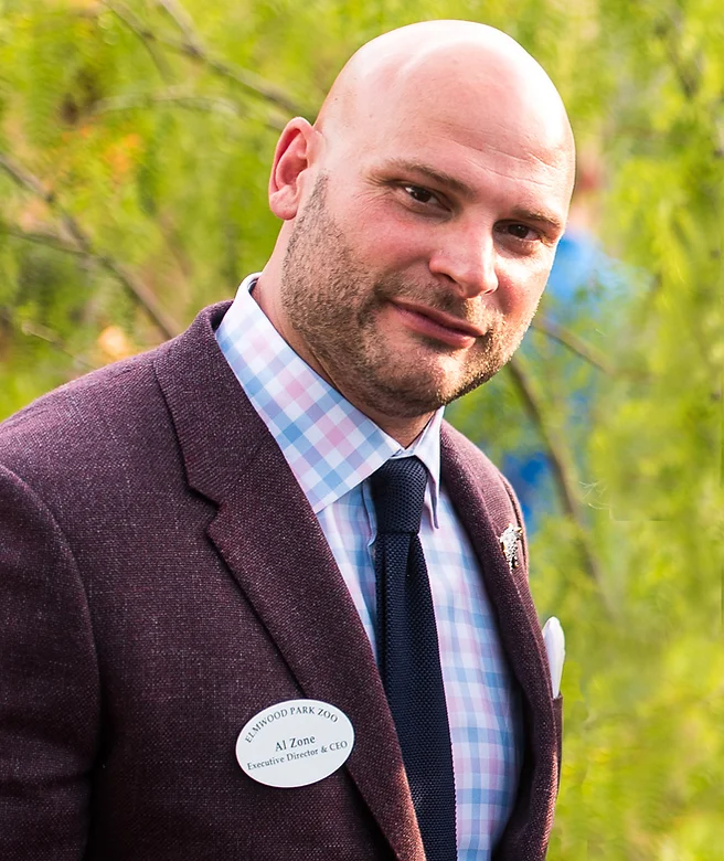 A bald man wearing a checkered dress shirt, a dark tie, and a maroon blazer with a name tag that reads Ellicwood Park Zoo, AI Zone, Executive Director & CEO, standing outdoors with green trees in the background.