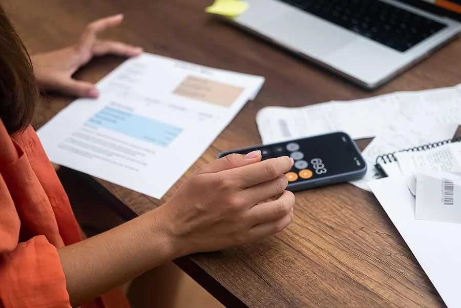 Person using a calculator on a wooden desk with scattered papers and a laptop.