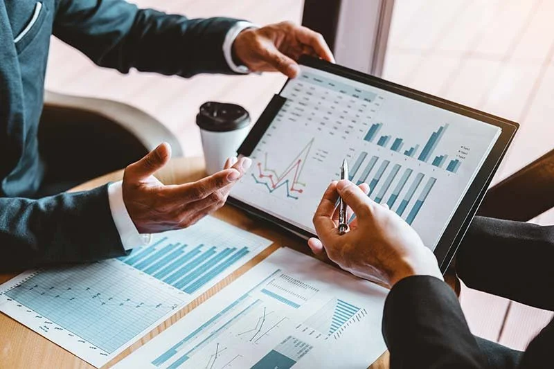 Two people in business attire analyzing financial data on a tablet and paper documents with charts and graphs in a conference room.