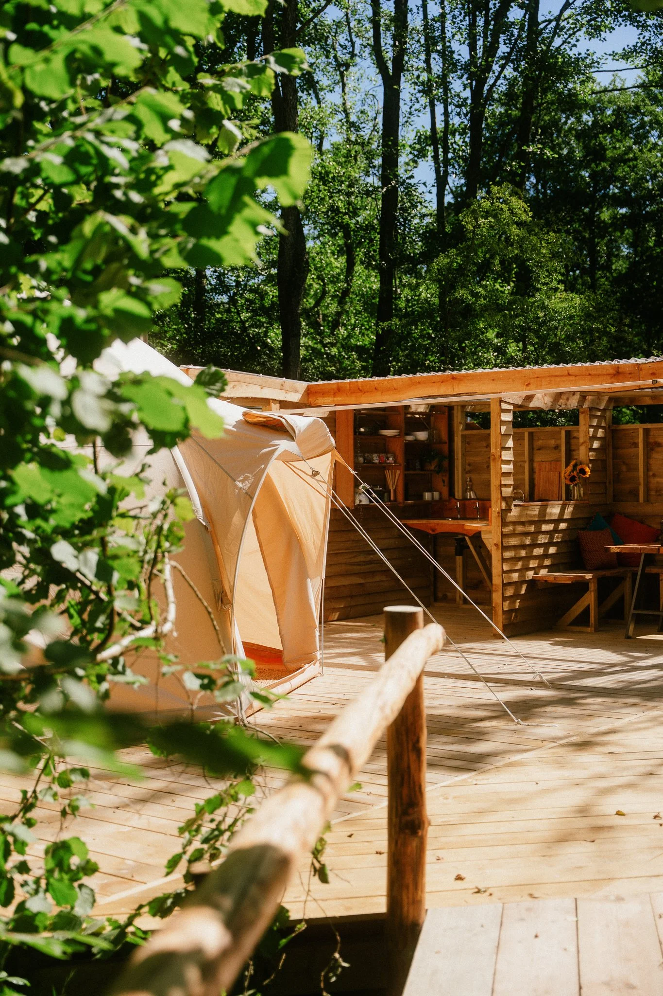 A wooden deck area surrounded by trees with a beige tent set up on the deck and a wooden outdoor kitchen area with shelves, a sink, and a sunflower in a vase.