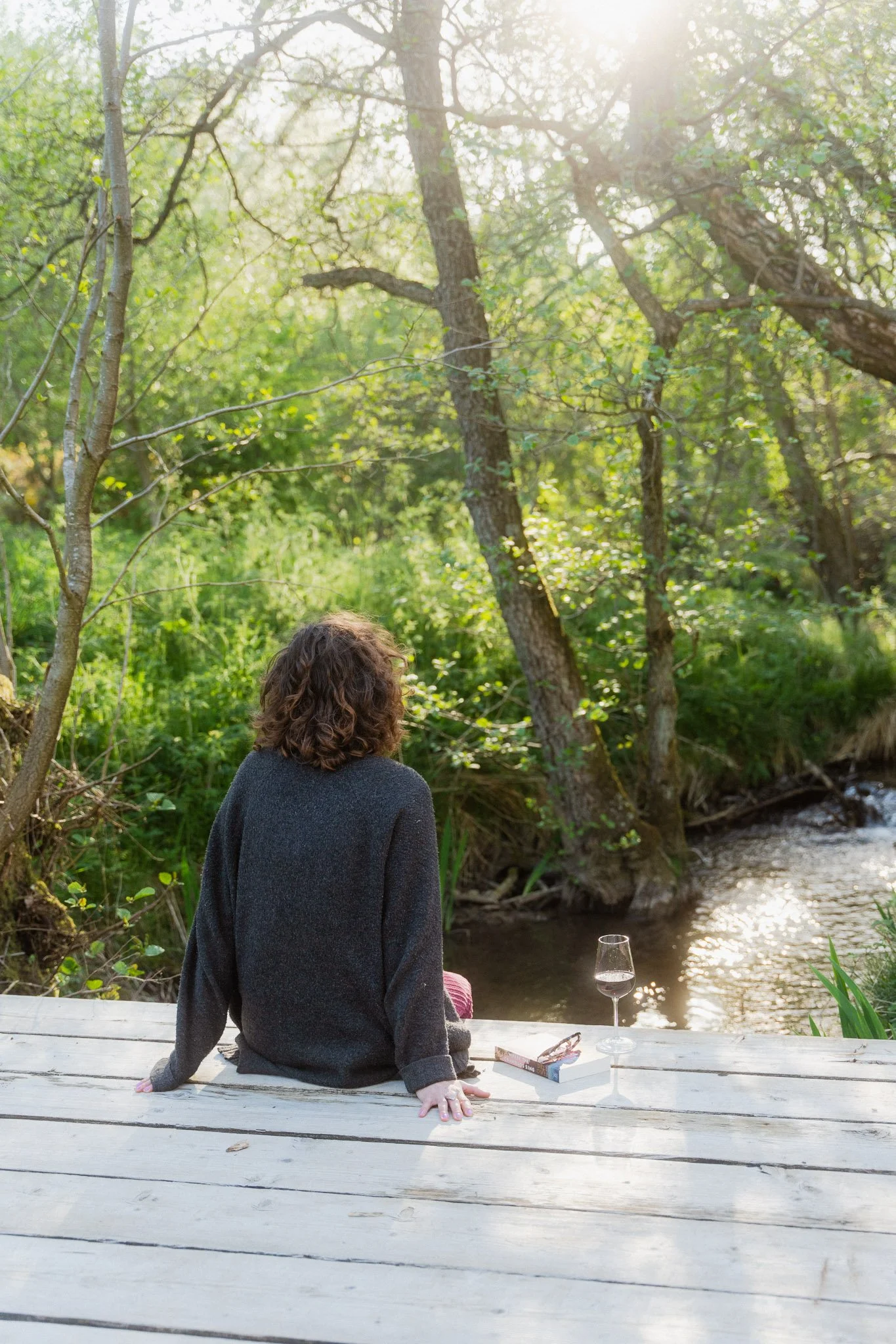 A person with curly hair sitting on a wooden dock by a stream, with a glass of wine and a pack of cigarettes nearby, surrounded by greenery and trees, with sunlight filtering through the leaves.