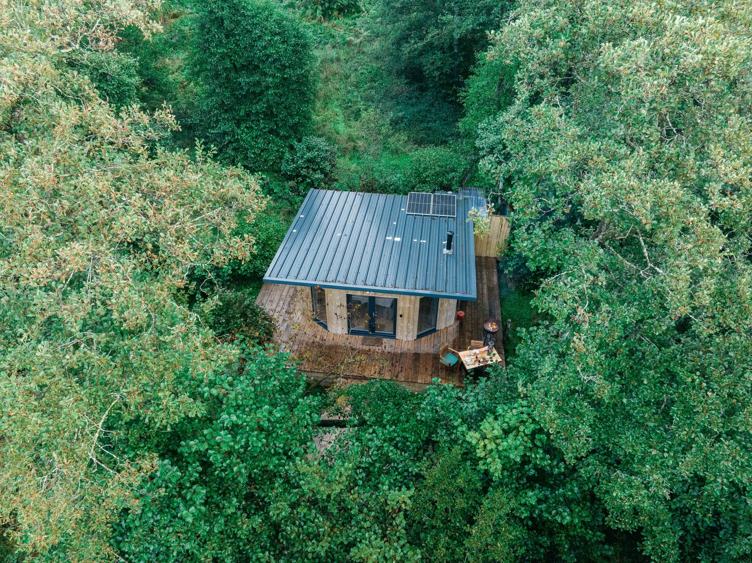 Aerial view of a small cabin surrounded by dense green trees with a deck that has outdoor furniture and a grill.