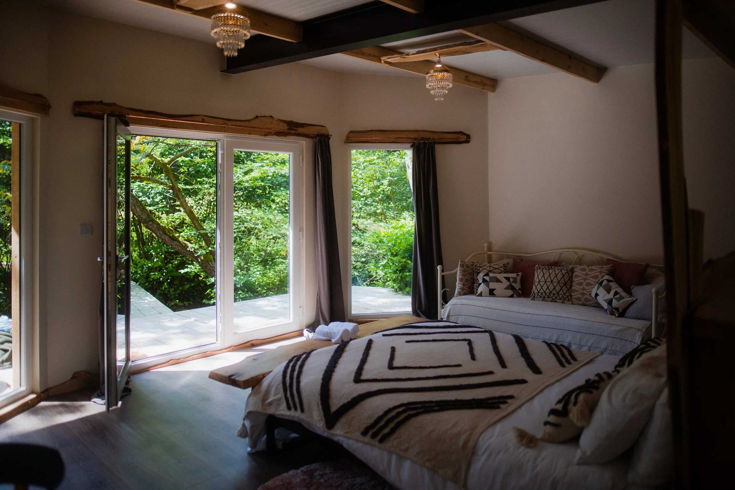 A cozy bedroom with sliding glass doors leading to a lush green outdoor area. The room features a bed with a beige and black patterned blanket and multiple decorative pillows, and a white daybed along the wall with similar pillows. The ceiling has wo