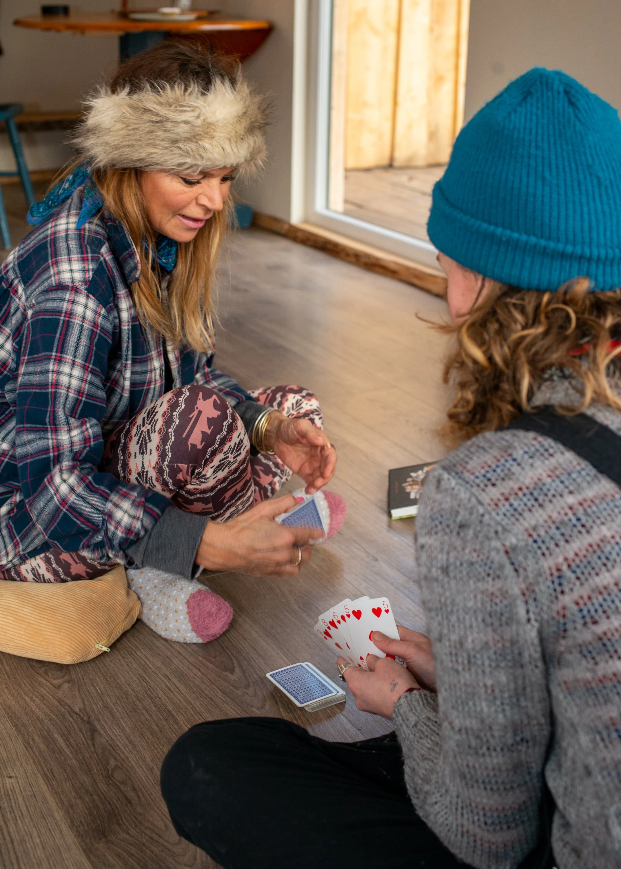 Two women sitting on a wood floor playing a card game indoors. One woman is wearing a furry hat, plaid shirt, and patterned pants, and the other is wearing a blue beanie and a gray sweater. There are cards and a small notebook on the floor.