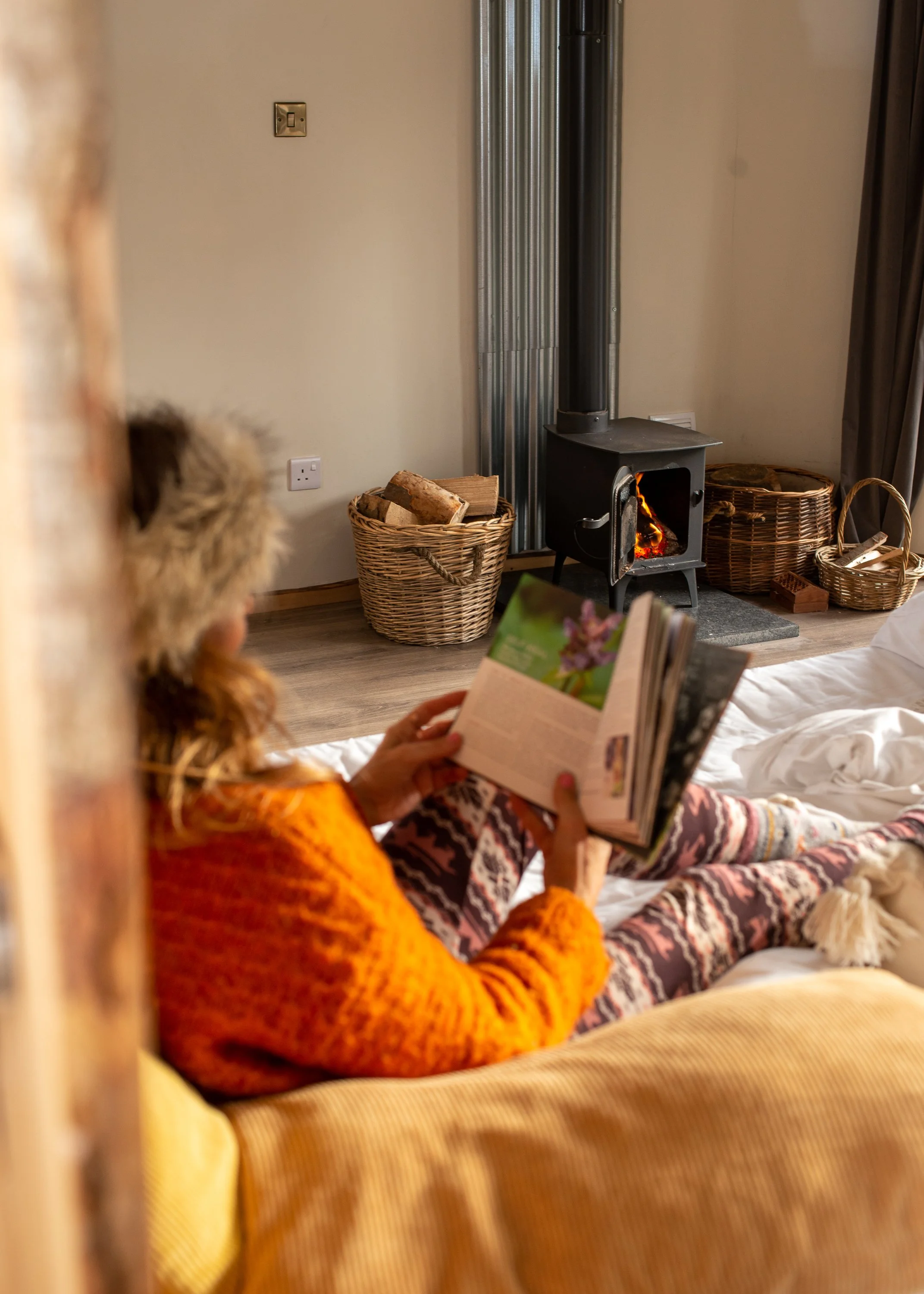 Person with long hair, wearing an orange sweater, sitting on a bed and reading a book, with a wood stove burning in the background.