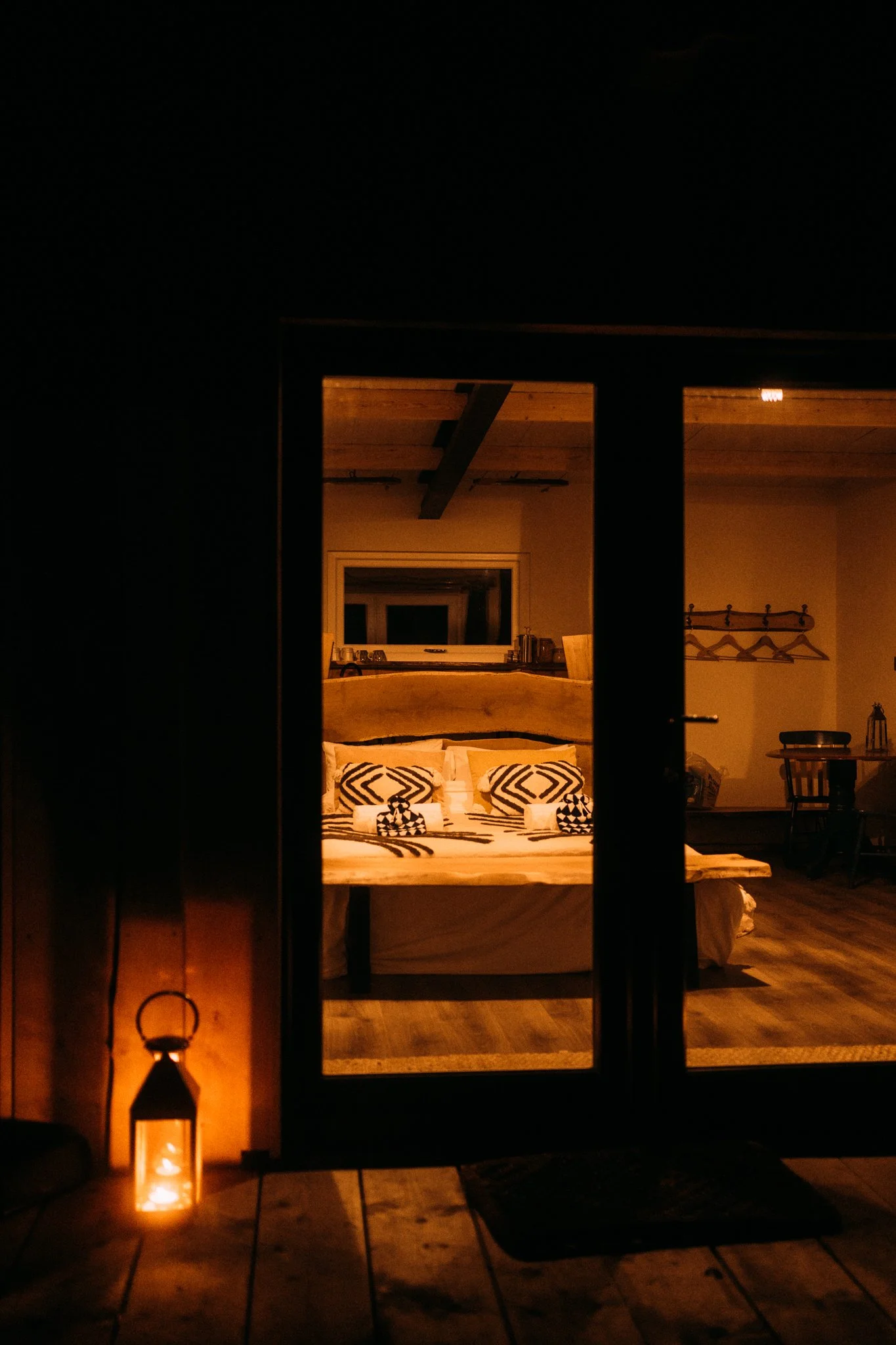 Cozy bedroom seen through glass doors, decorated with patterned pillows, warm lighting, and wooden furniture.