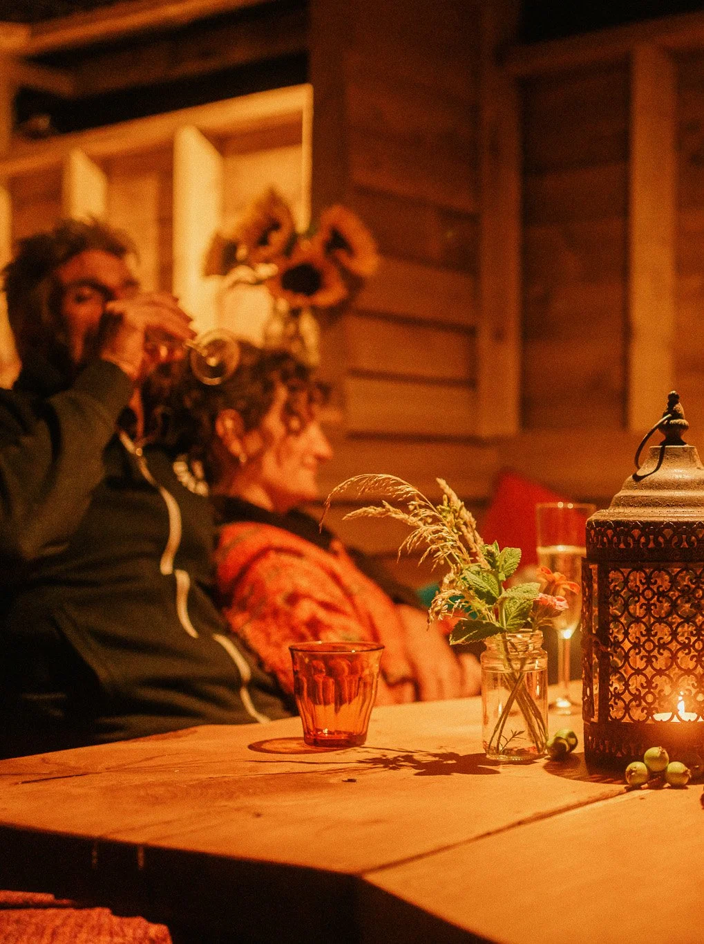 People sitting at a wooden table in a dimly lit cozy room, with a lantern, glass, and vase with flowers for decoration.