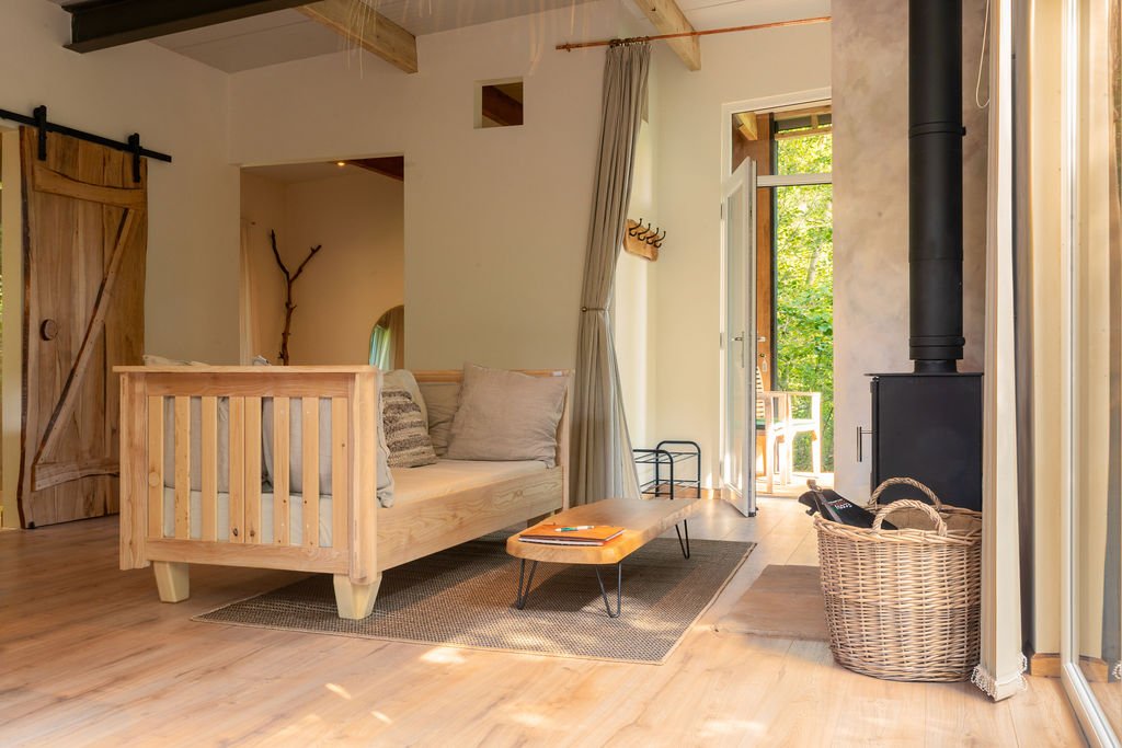 Cosy wabi-sabi living room with custom made wooden furniture, a wood stove, and a balcony door opening to green outdoors.