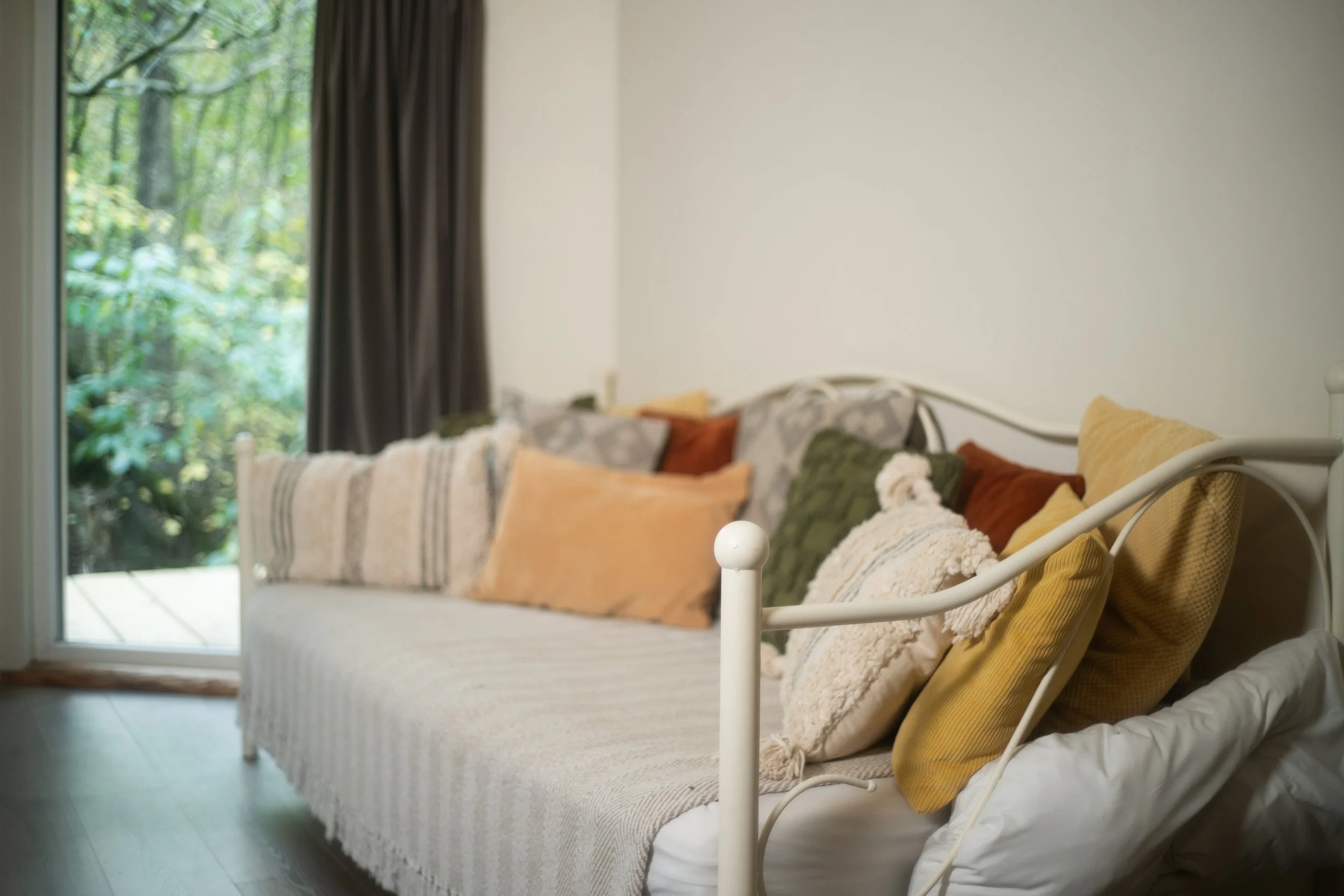 A cosy living room with a white metal-framed sofa layered with various colored pillows, near a glass door showing greenery outside. Optional single bed at Brook