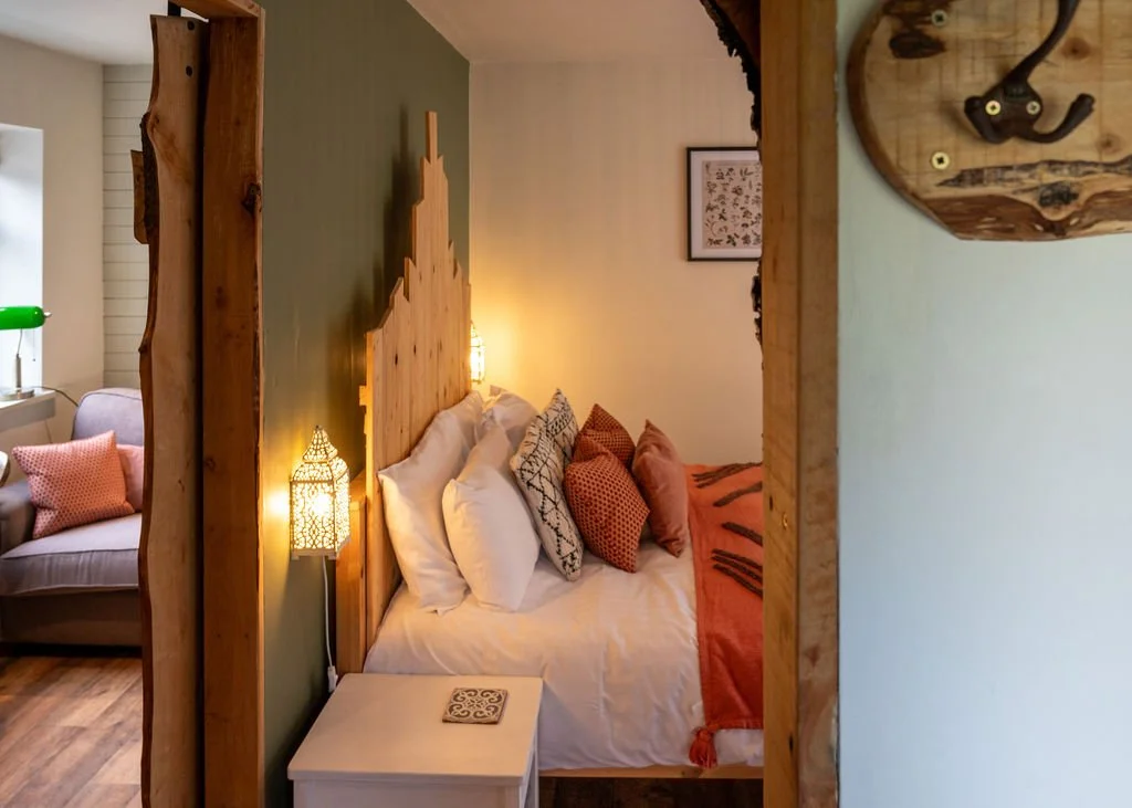 A cozy bedroom with a wooden headboard and colorful pillows on a bed, a side table, and warm lighting. Adjacent seating area with a grey sofa and pink cushions is visible in the background.