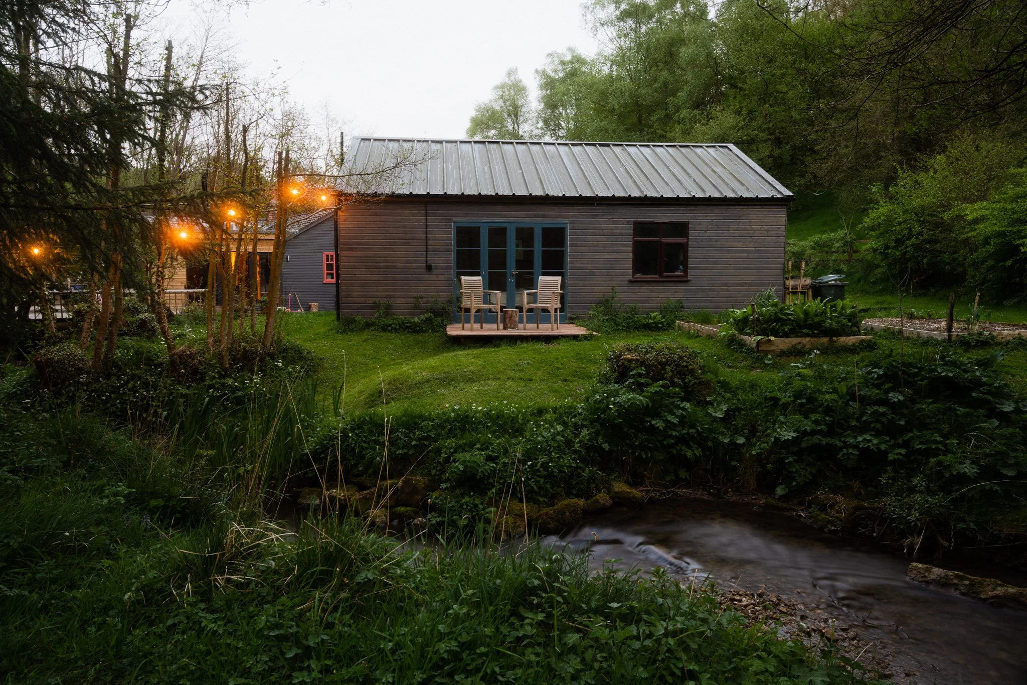 A small wooden house with a metal roof, surrounded by greenery, with a small porch and outdoor chairs. String lights hang from nearby trees. There is a creek in the foreground and a garden bed on the right side.