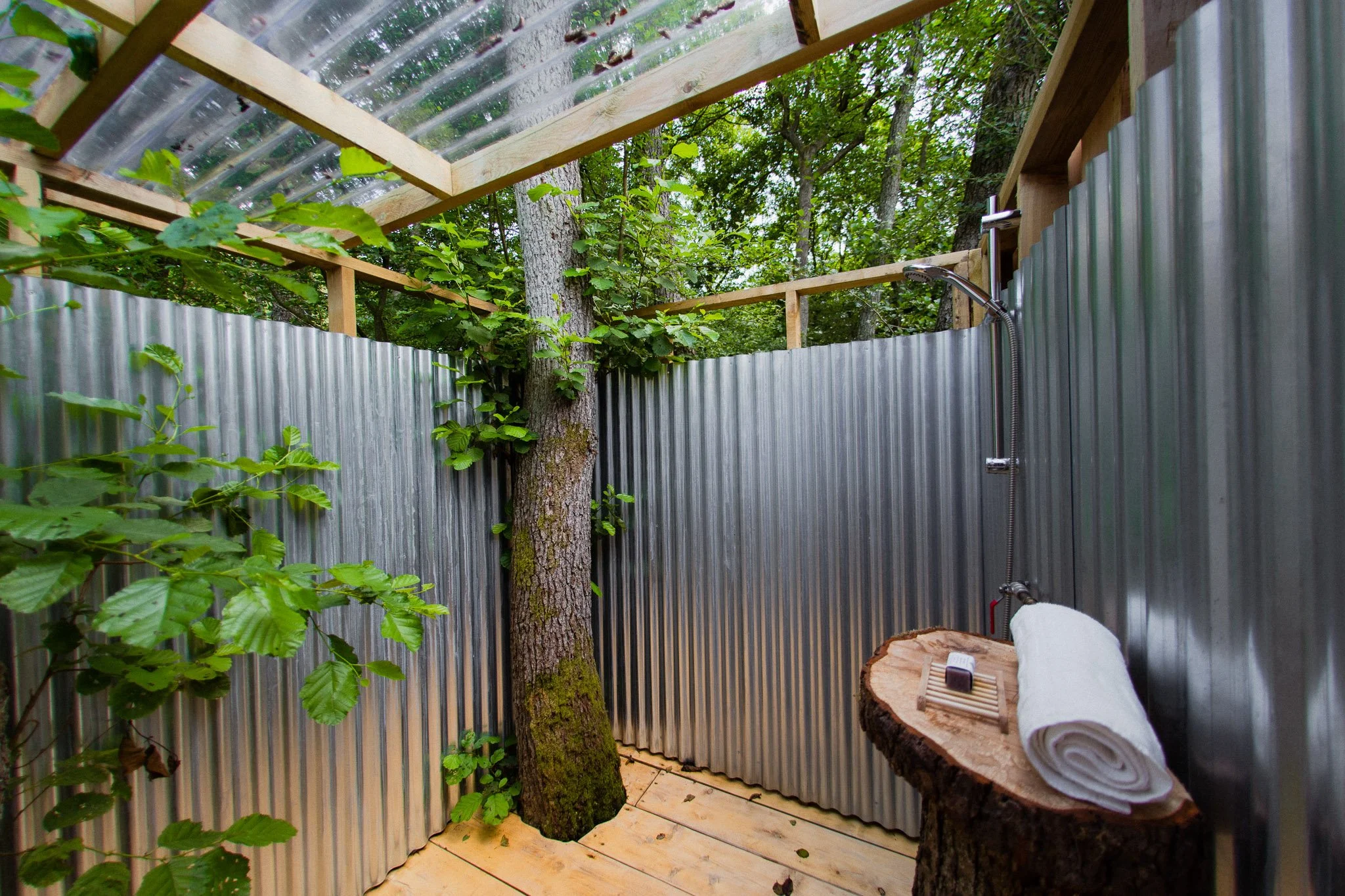 An outdoor shower area with metal corrugated walls, wooden flooring, and a tree growing inside. There is a small wooden shelf with a rolled towel and shower accessories on a tree stump.