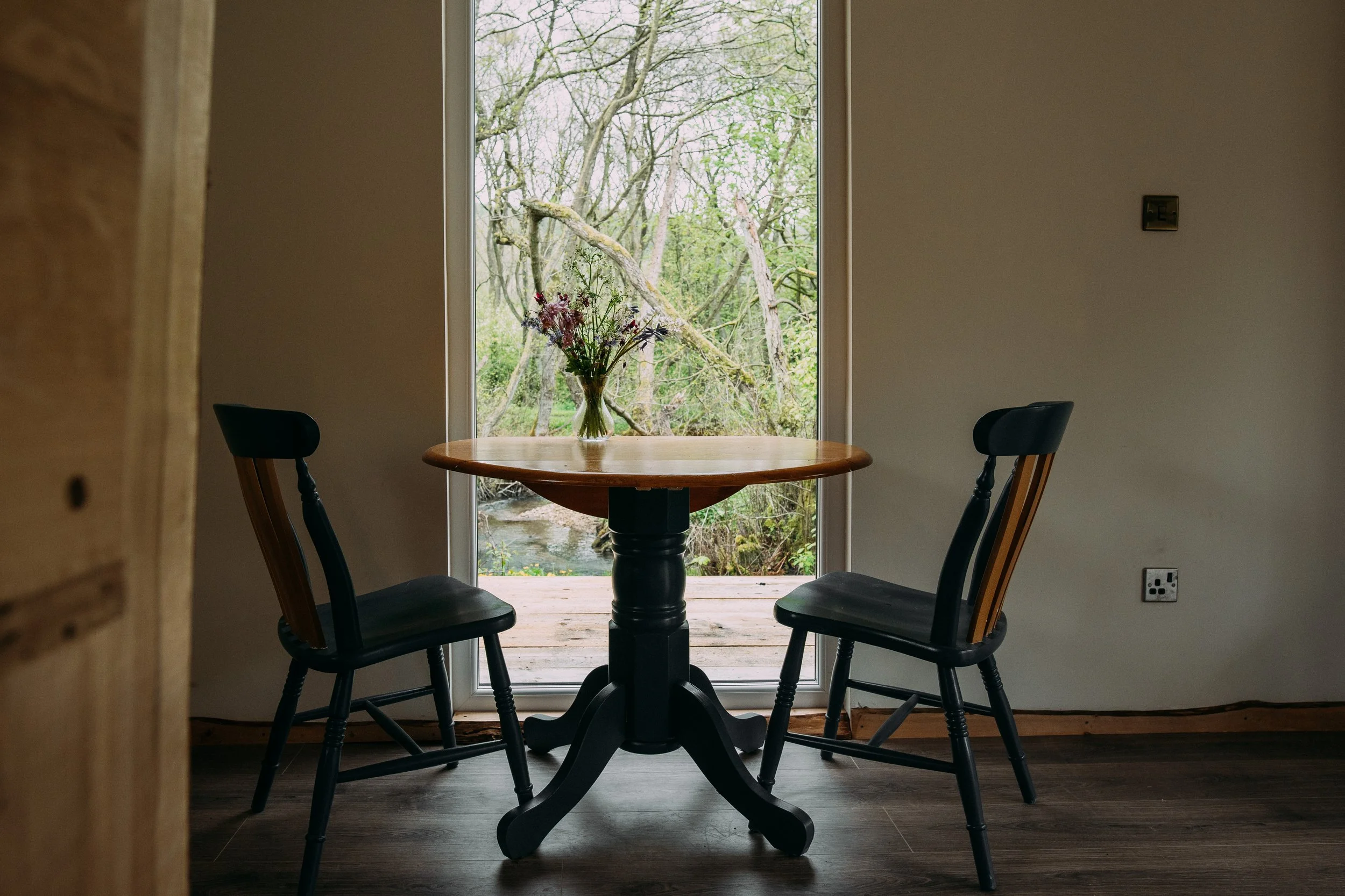 A round wooden table with a glass vase of flowers, two black wooden chairs, and a large window showing trees outside, breakfast view at Brook